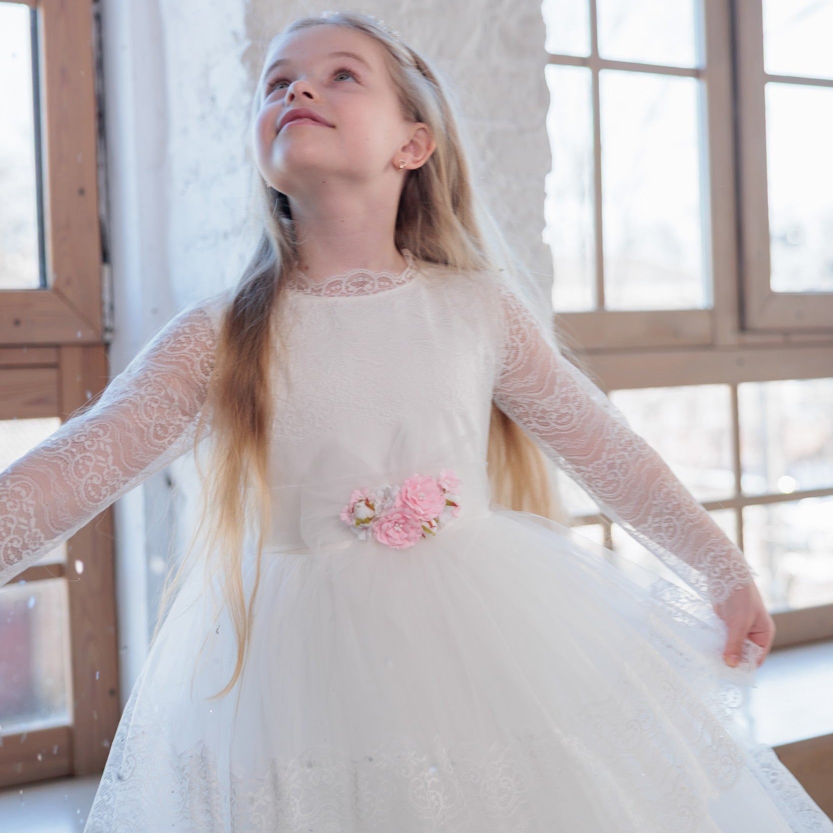 Young girl in an ivory lace communion dress with floral details standing in front of large windows.