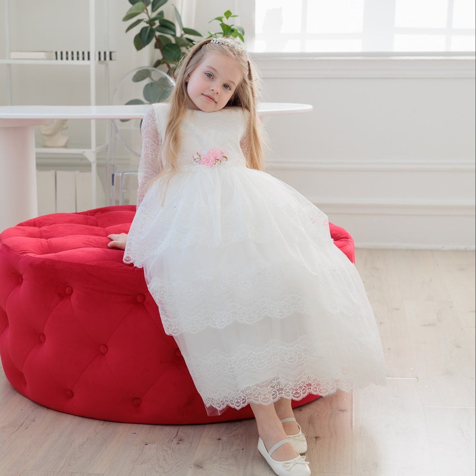 Young girl in a white dress sitting on a red bean bag chair in a bright room.