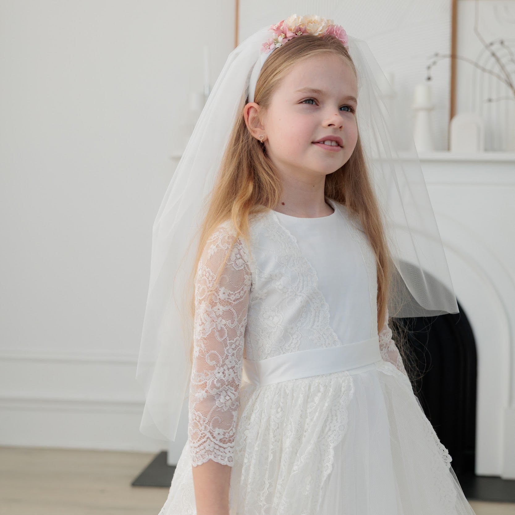Young girl in an ivory lace communion dress  with lace sleeves and a veil indoors.