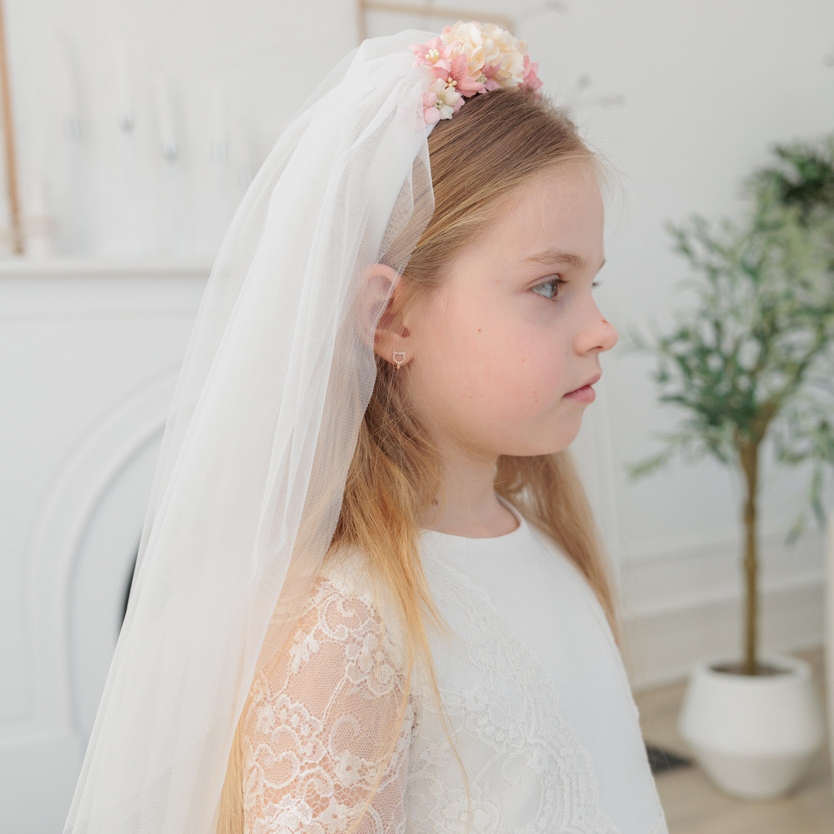 Young girl in an ivory lace communion dress with a lace veil and floral headband indoors.