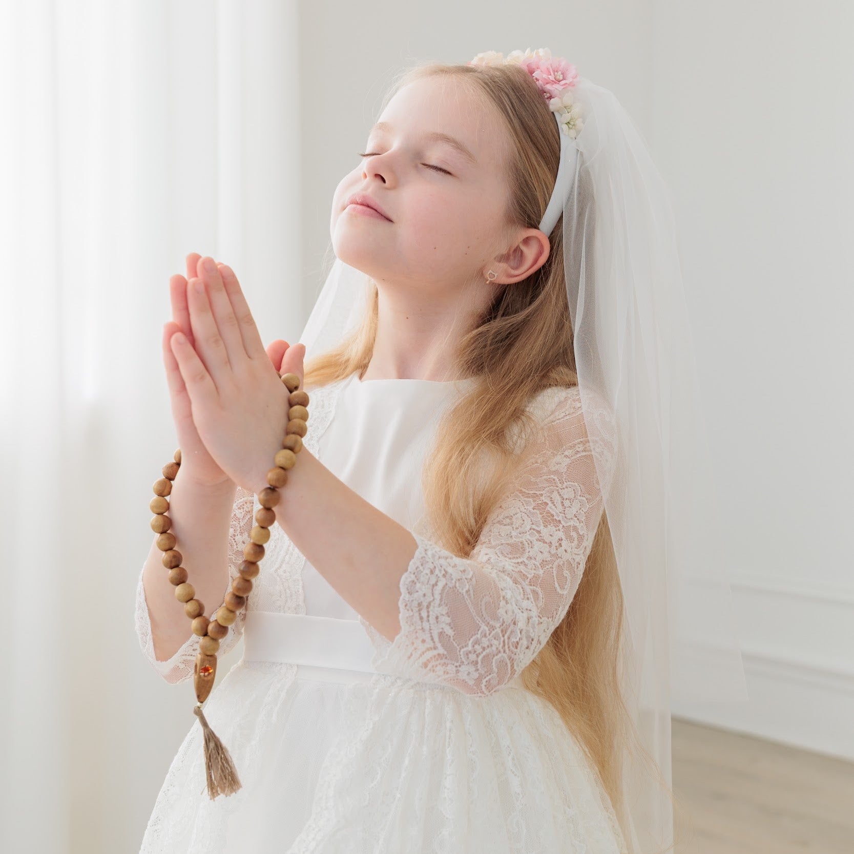 Young girl in an ivory lace communion dress  with lace sleeves holding prayer beads against a white background