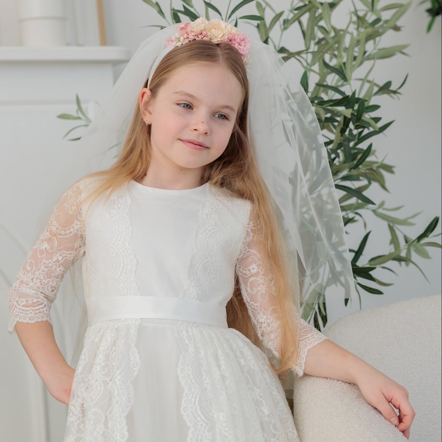 Young girl in an ivory lace communion dress  with a veil sitting on a couch.