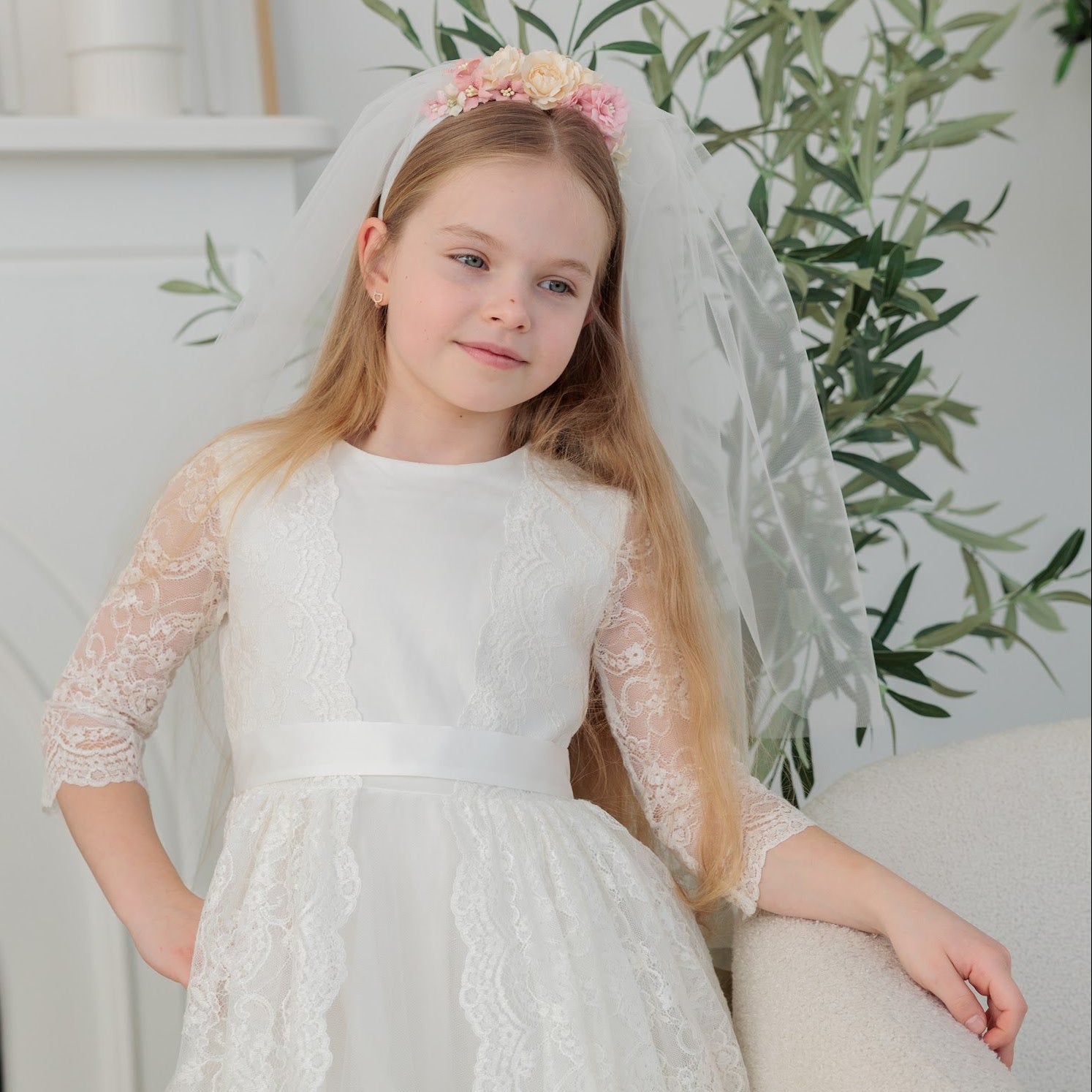 Young girl in a white lace communion dress with a communion veil sitting on a couch.