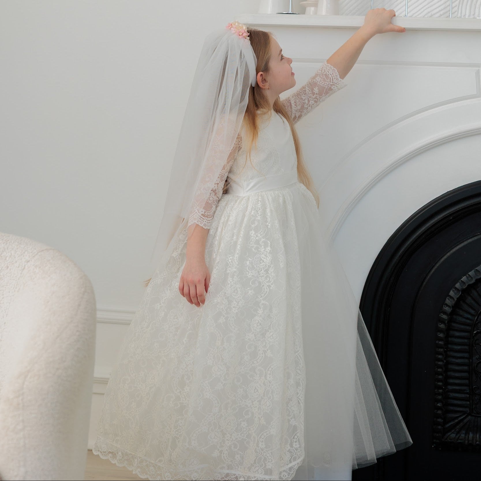 Woman in a white lace dress standing in front of a fireplace.