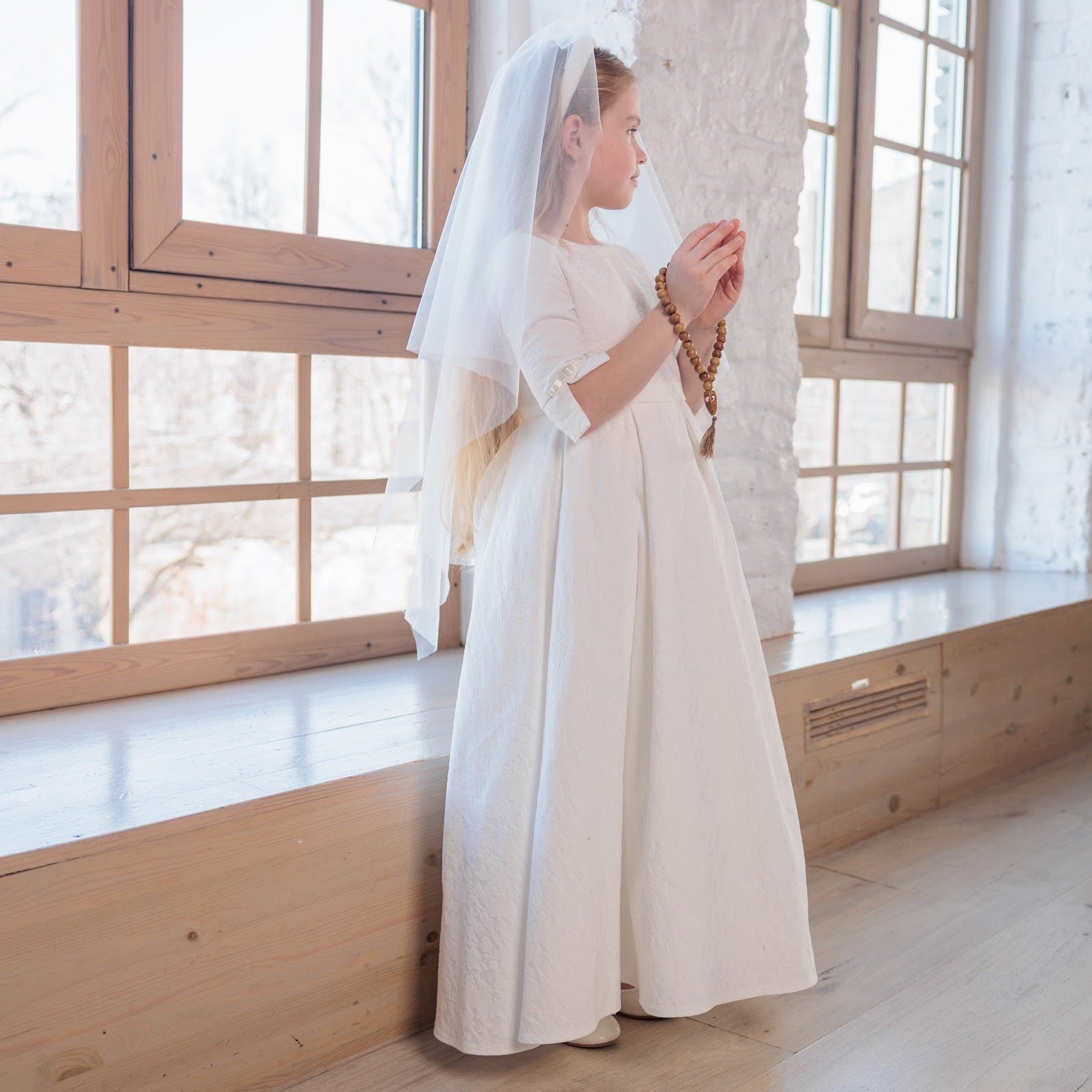 Woman in a white communion dress standing in a room with large windows.