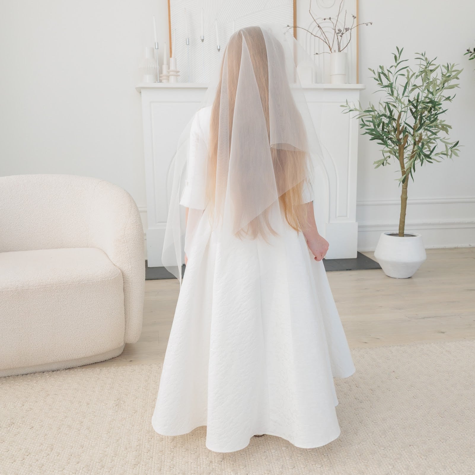 Person wearing a white communion dress with a long veil in a minimalistic room.