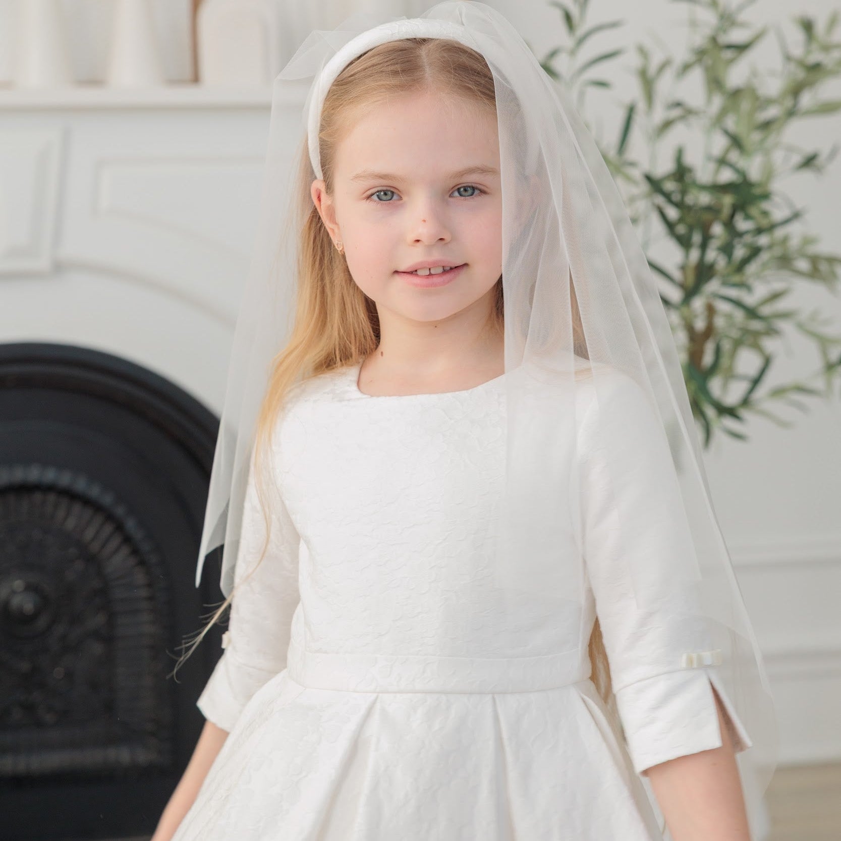 Young girl in a white communion dress with a veil standing in a room with a fireplace and plant.
