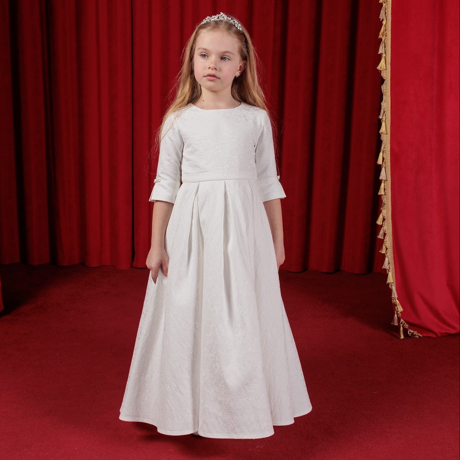 Young girl in a white communion dress standing in front of red curtains