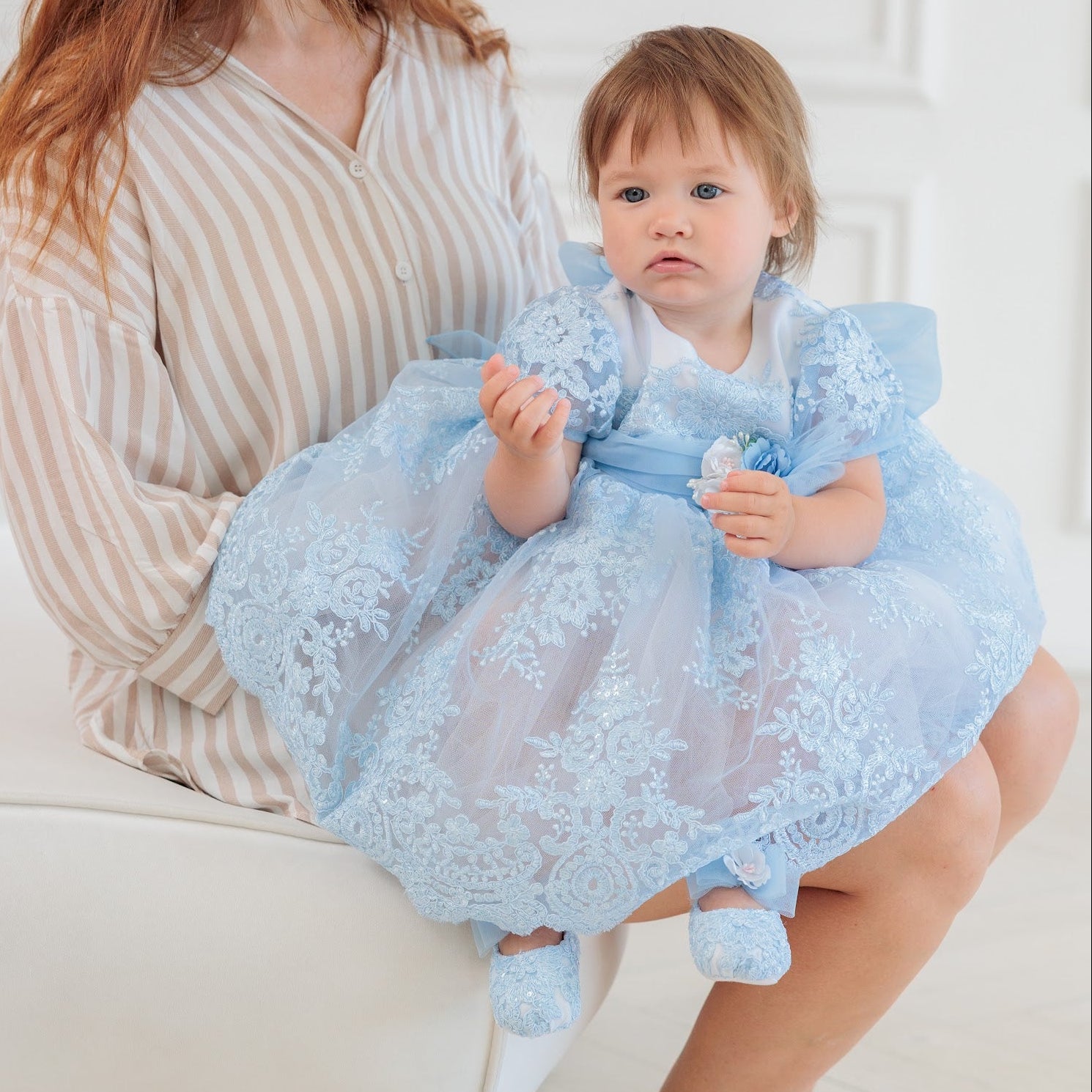 A woman sits on a white bench holding a toddler wearing a light blue lace dress with matching blue booties in a bright indoor room.