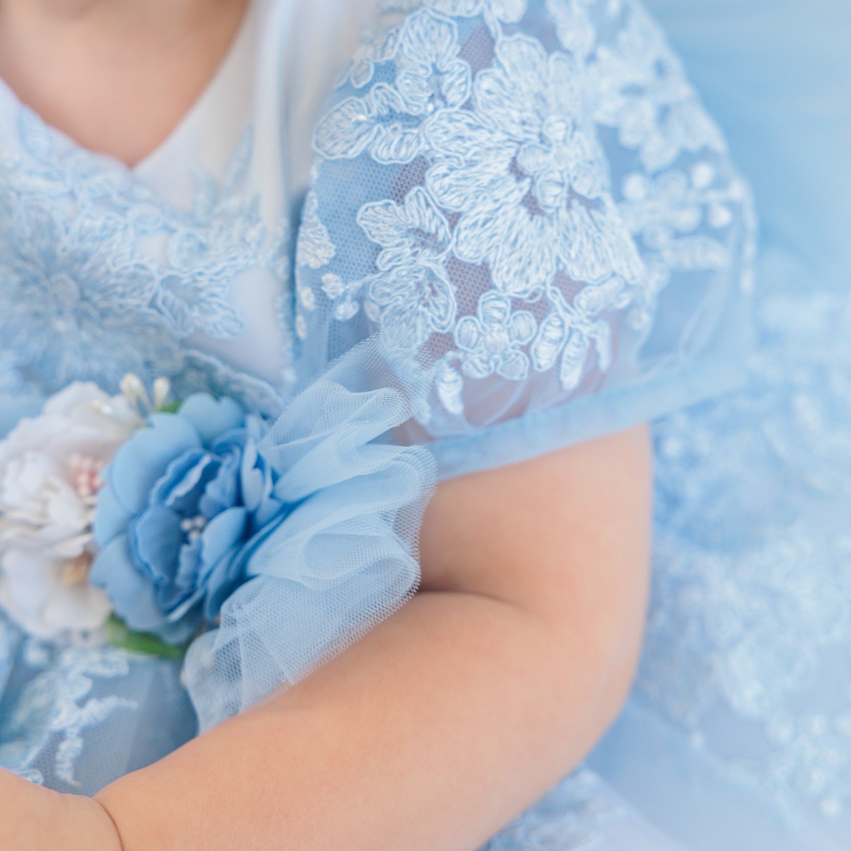 A close-up of a child wearing a light blue lace dress with floral embroidery, sheer tulle sleeves, and a decorative blue fabric flower accessory on the sleeve.