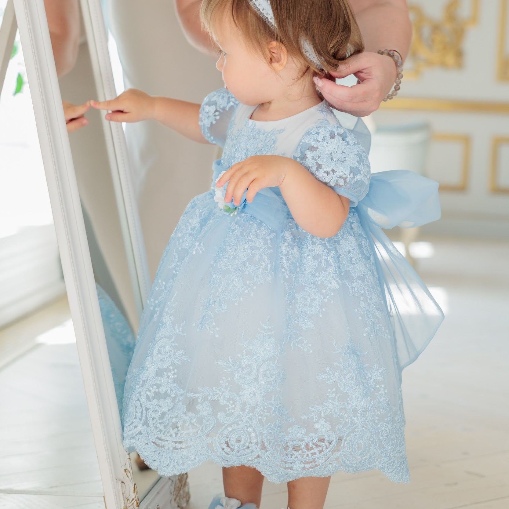 A toddler girl wearing a light blue lace and tulle dress with a bow, standing in front of a mirror while an adult adjusts her headband in a bright indoor room.
