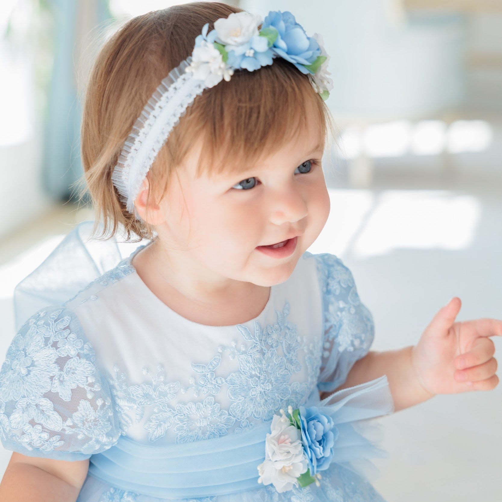 A toddler girl wearing a light blue lace dress with a matching blue and white floral headband, pointing toward the right in a bright indoor setting.