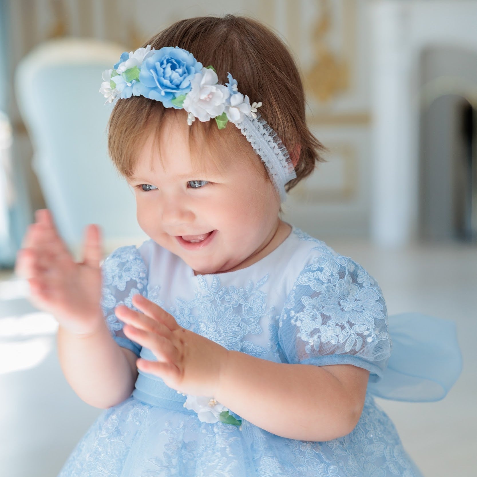 A smiling toddler wearing a light blue lace dress and a matching blue and white floral headband, standing indoors with hands raised.