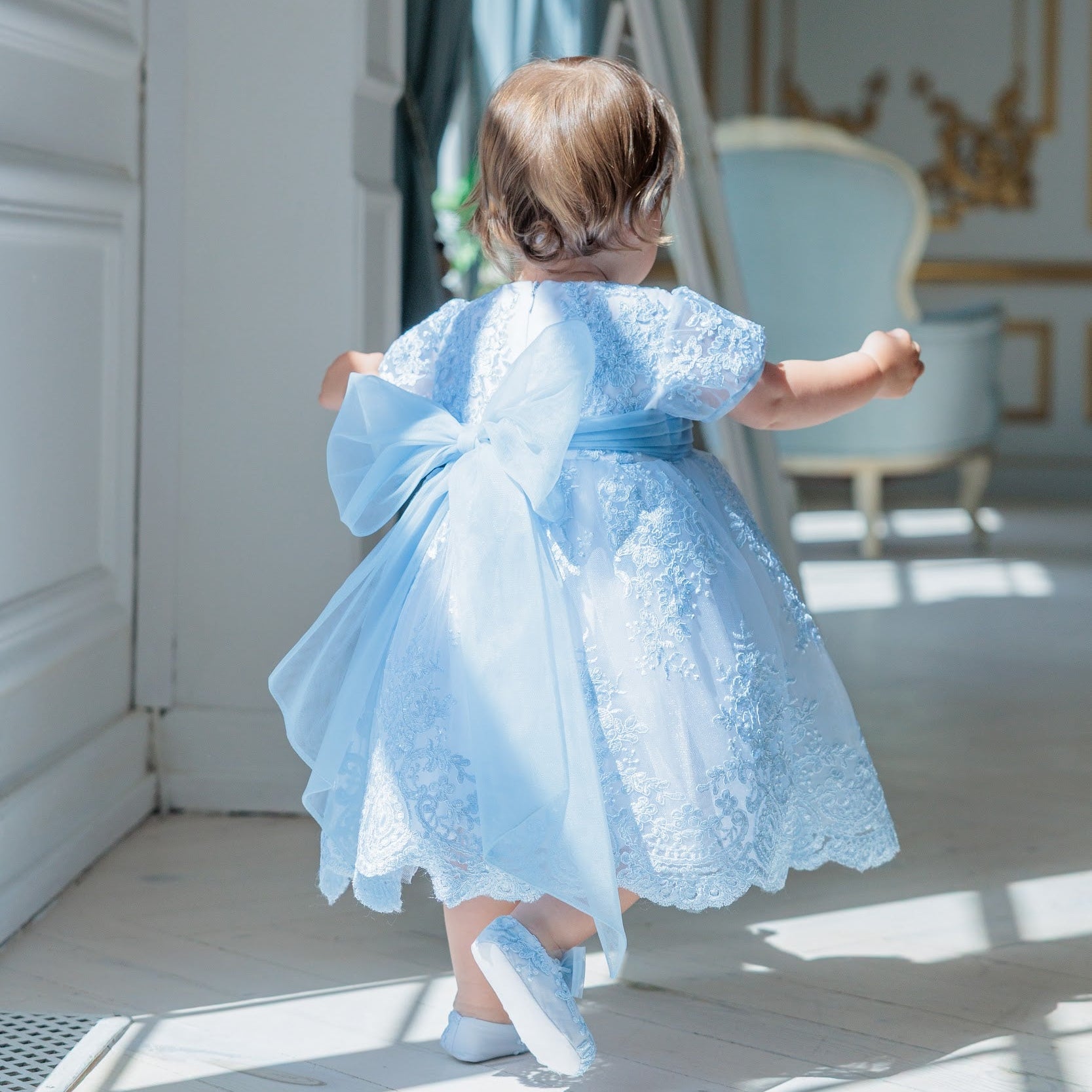 A toddler girl seen from behind wearing a light blue lace and tulle formal dress with a large bow on the back, standing in a bright indoor room.