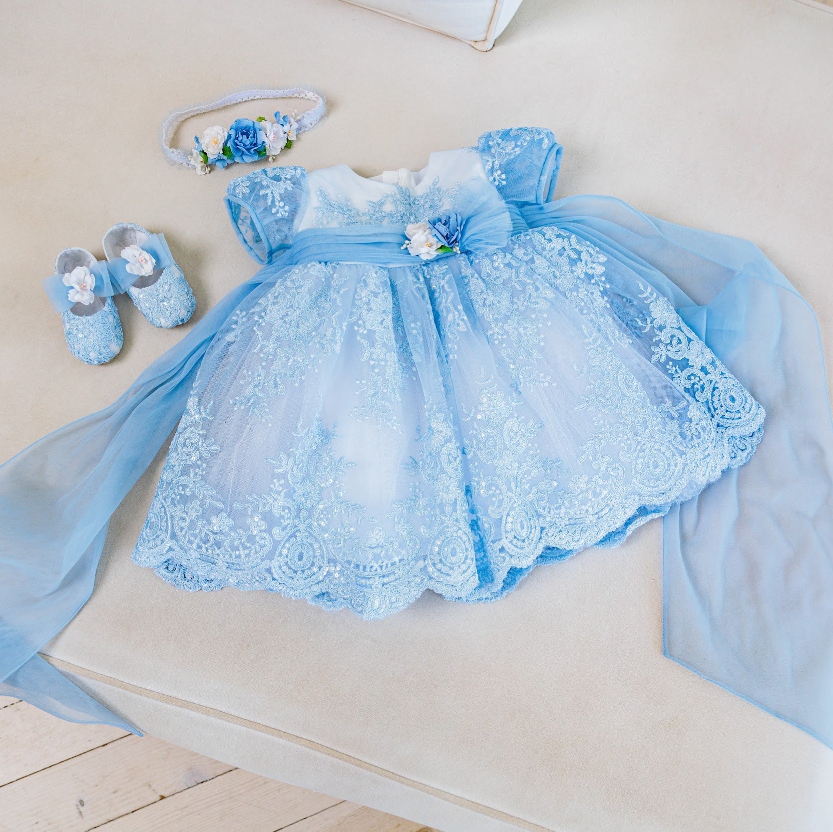 A baby girl's flower dress set in light blue lace and tulle, featuring a matching headband and blue booties, laid out on a beige upholstered bench.
