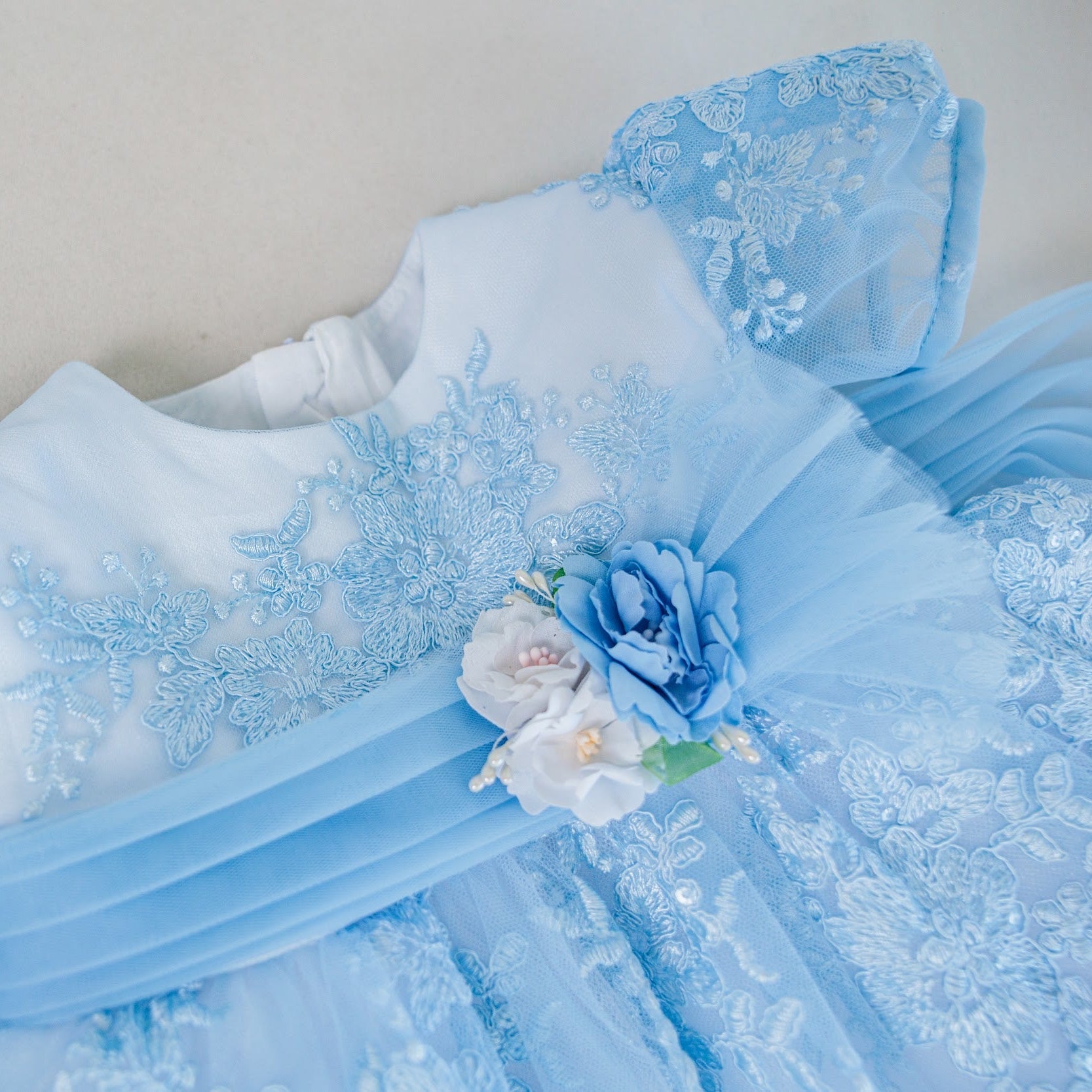 A close-up of a light blue lace and tulle garment laid flat on a neutral beige background, featuring floral applique, sheer mesh panels, and a decorative fabric flower accent.