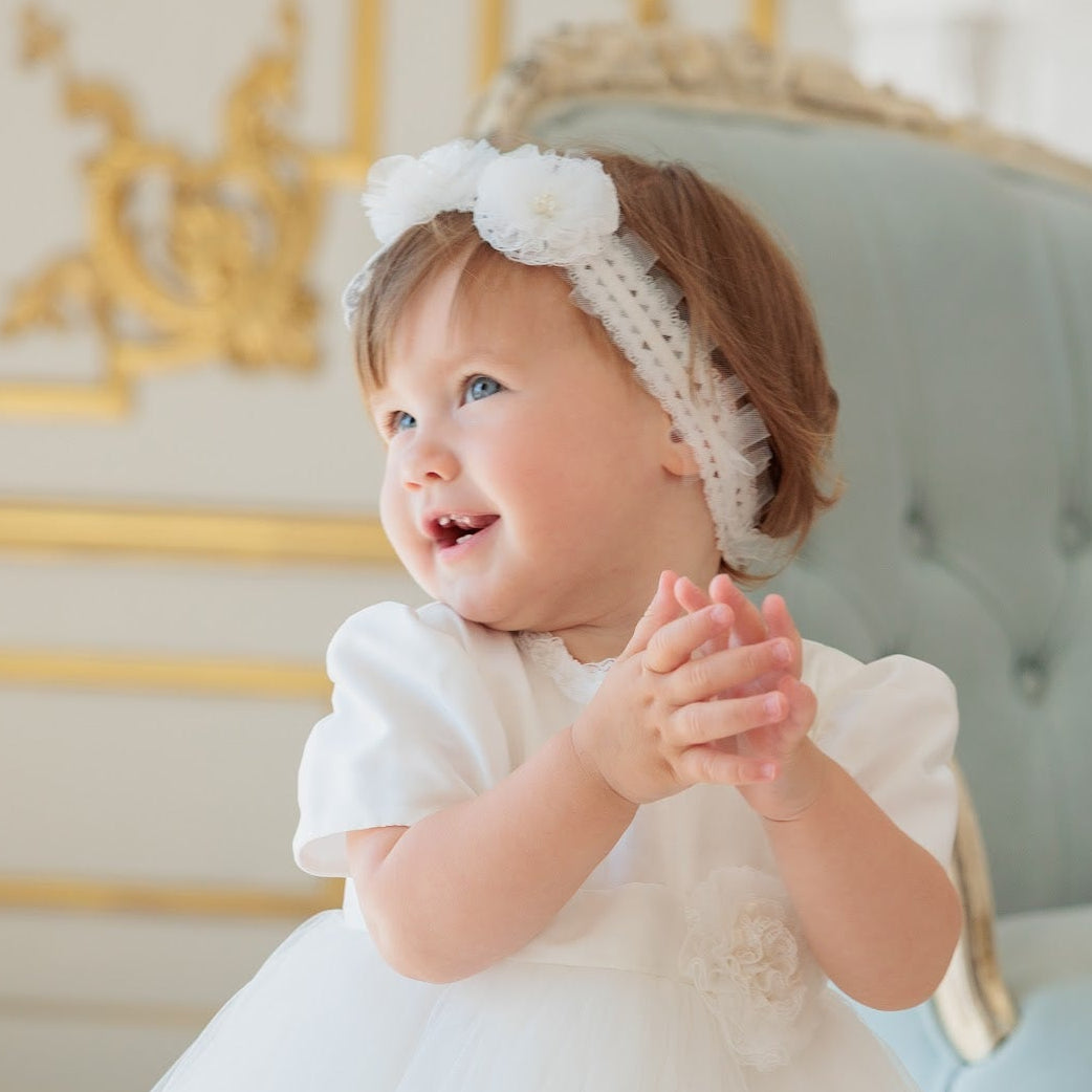 A smiling toddler girl wearing a white short-sleeve dress and a white floral lace headband, sitting in front of a light blue tufted chair with gold trim.