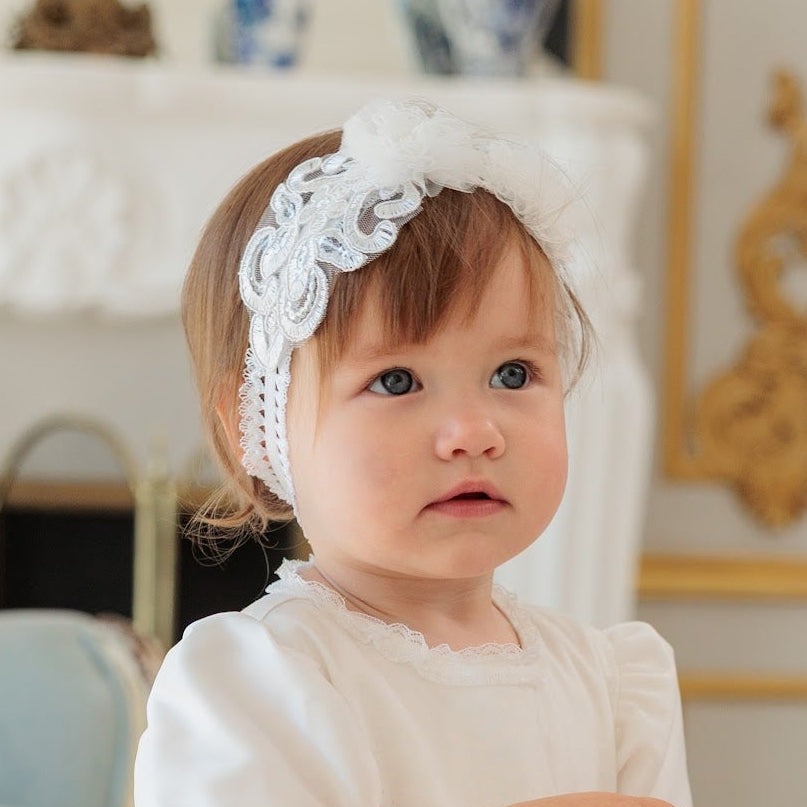 A toddler girl wearing a short-sleeved white lace dress with a matching white lace headband, standing in a bright, elegant room with gold-trimmed walls and light wood floors.