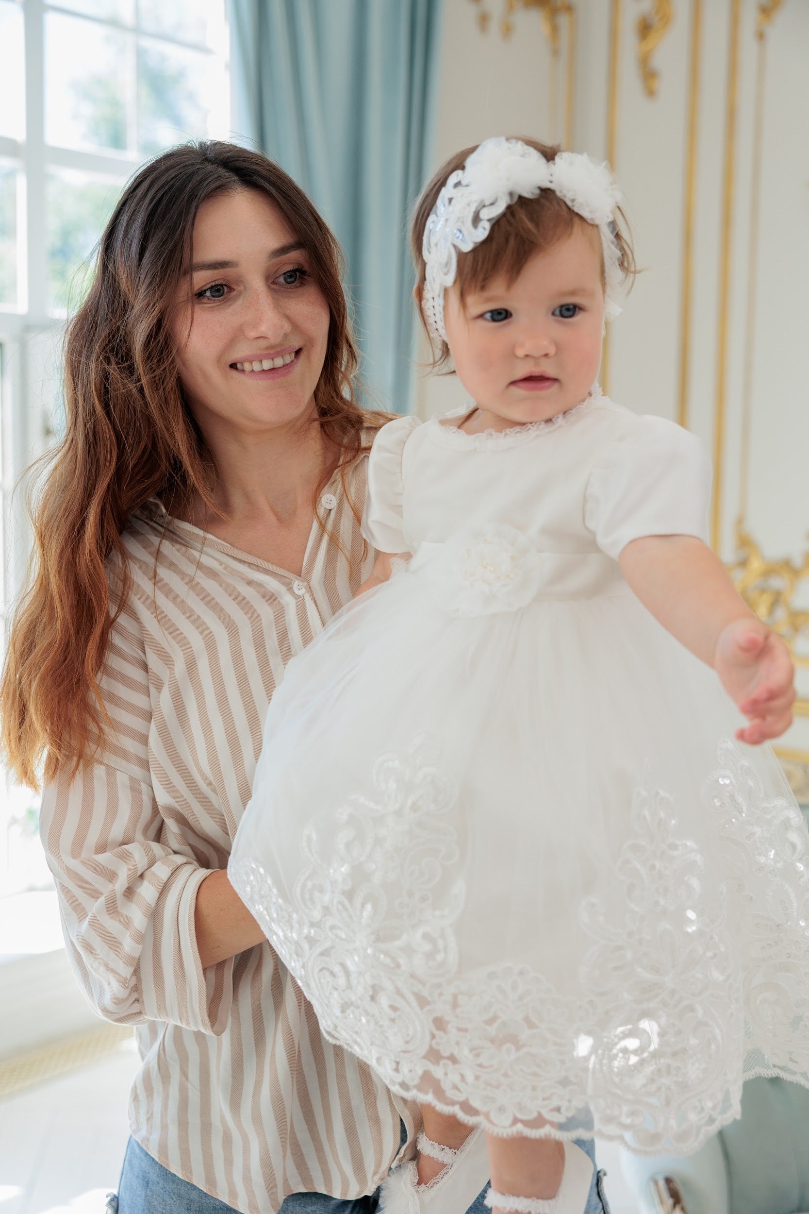 A woman holding a baby girl wearing a white lace first communion dress and matching white floral headband in an elegant indoor setting.