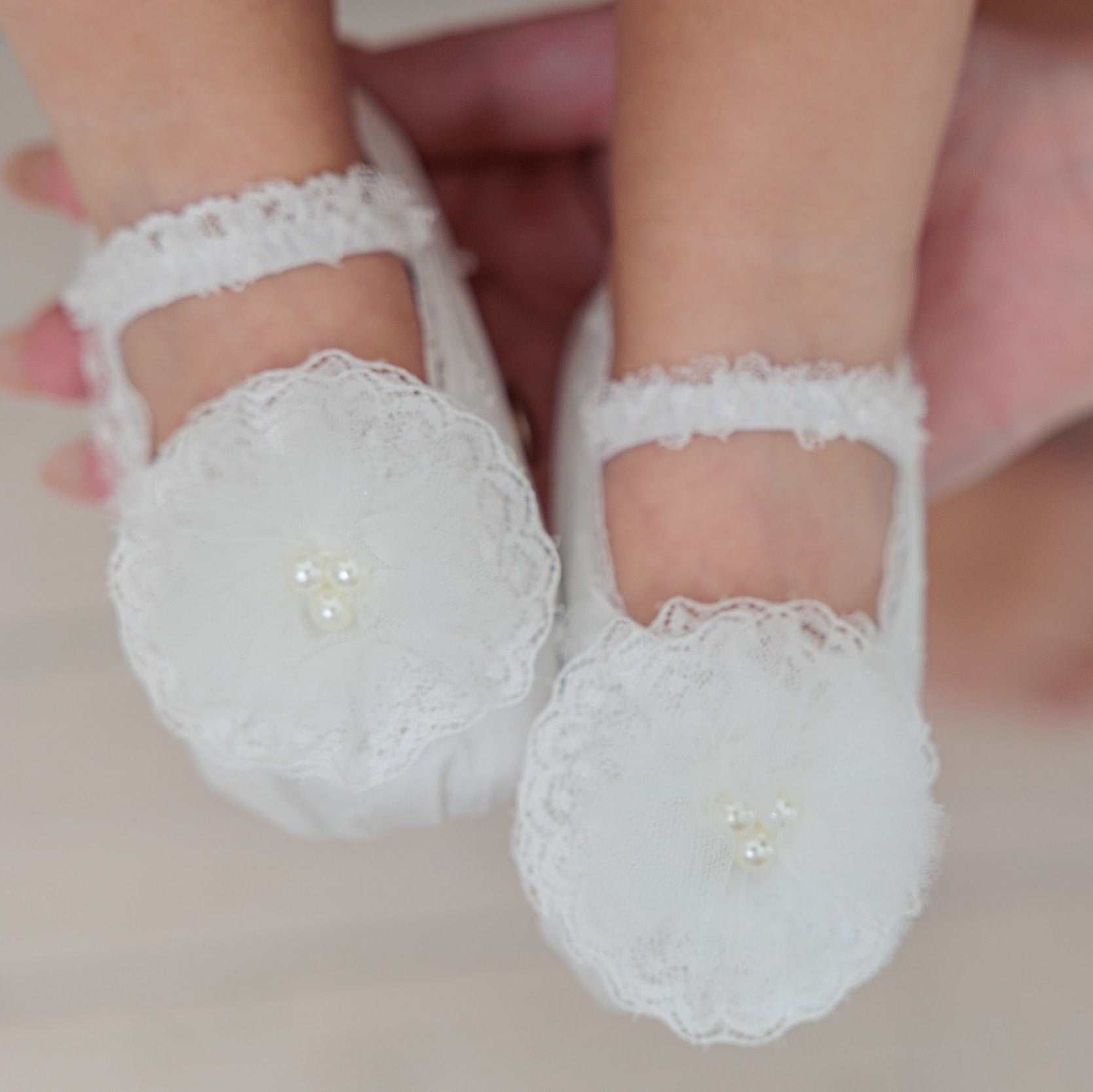 White lace baby booties with pearl embellishments, shown on a baby's feet held by an adult hand against a soft neutral background.