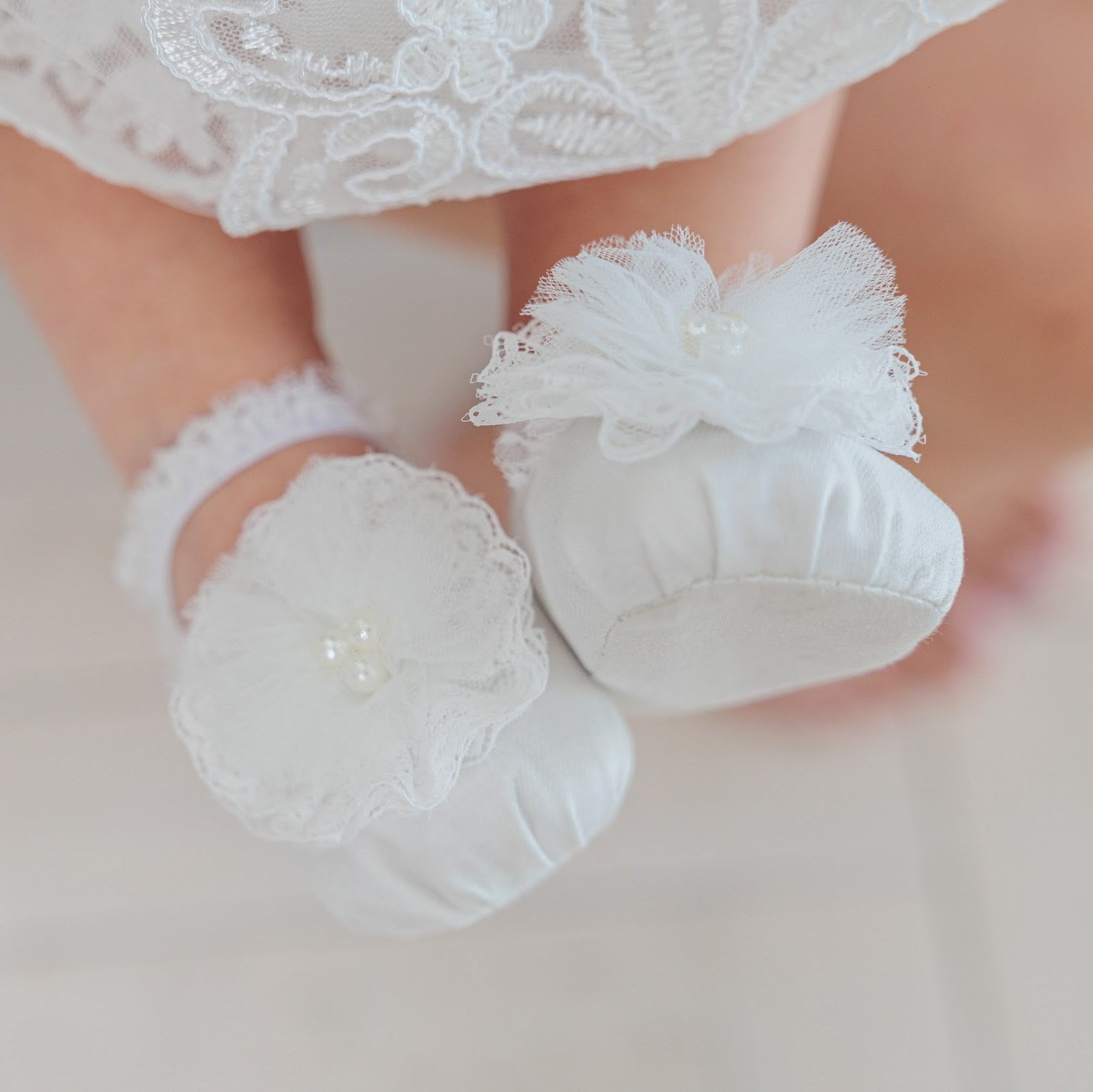 White lace baby booties with ruffled bows and pearl accents, shown on a baby's feet against a soft light background.