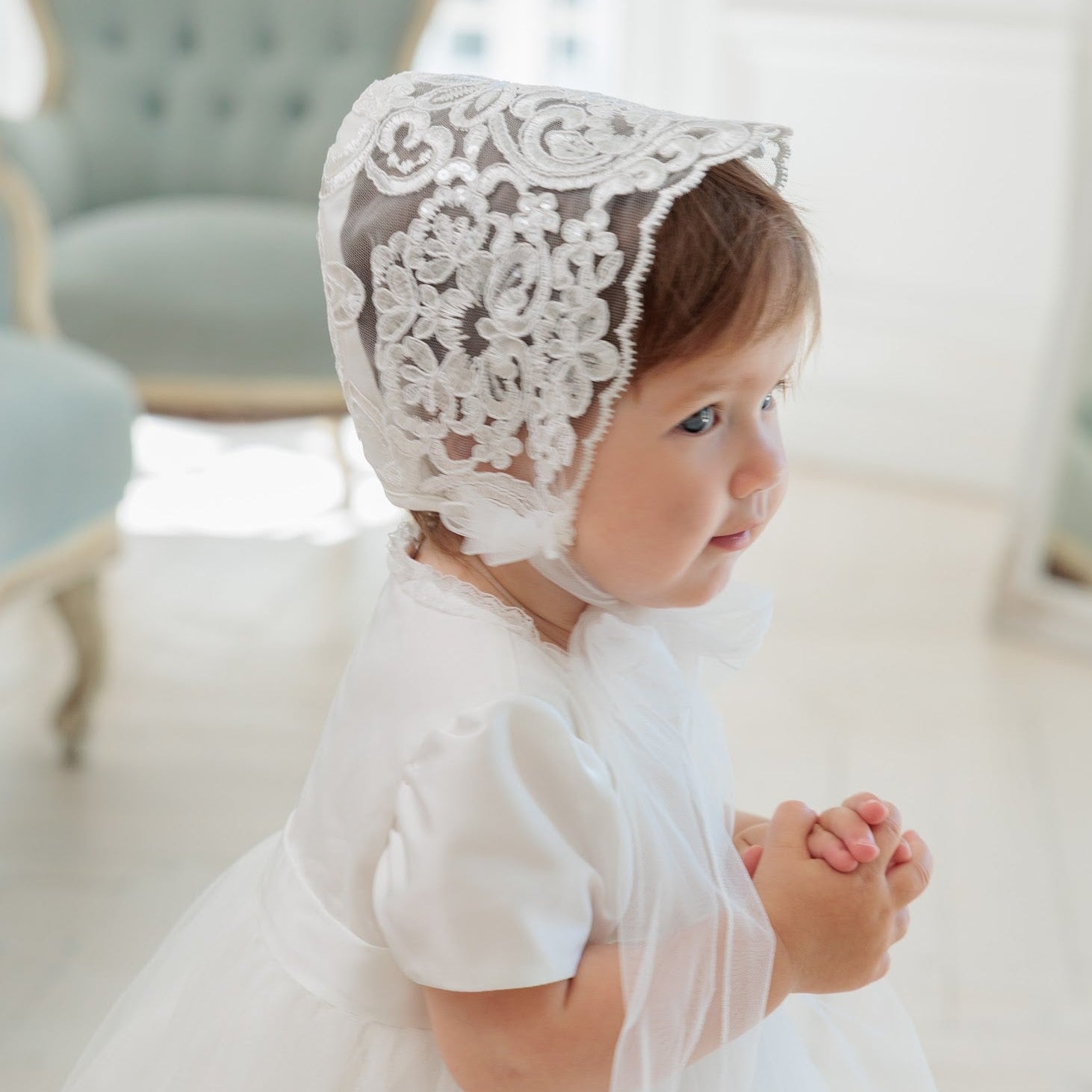 A toddler wearing a white dress and a white lace bonnet with floral embroidery, photographed in a bright indoor setting with a soft-focus blue chair in the background.