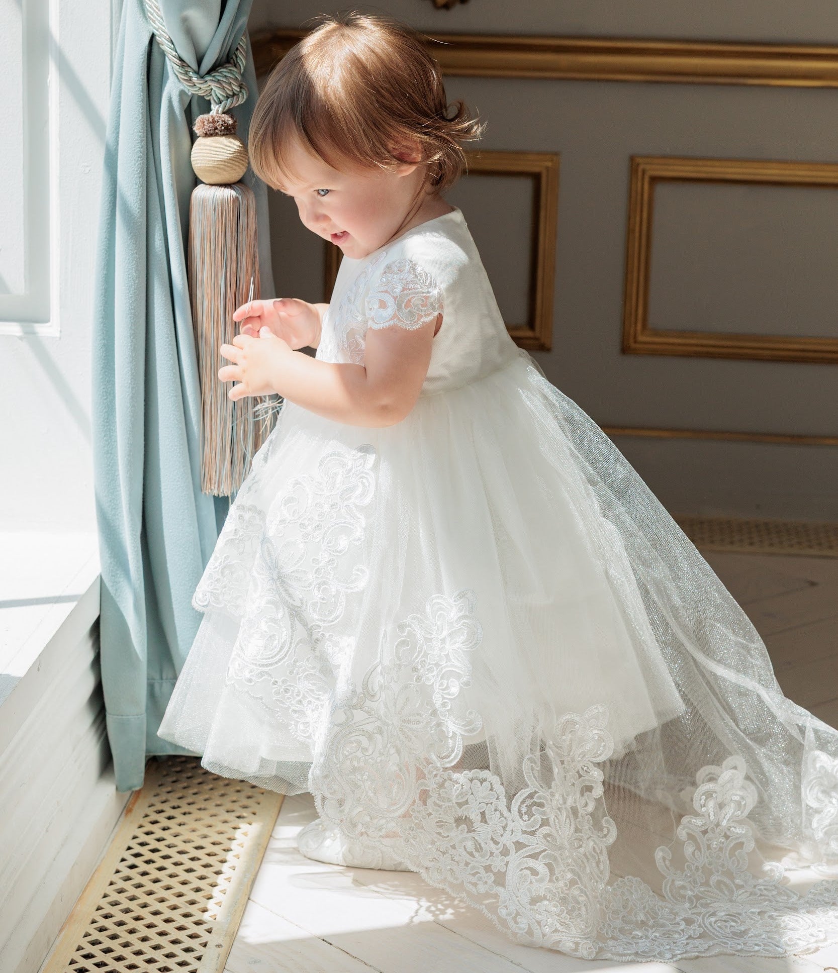A toddler girl wearing a white lace and tulle baptism dress stands by a window in an ornate room with gold-trimmed walls and blue curtains.