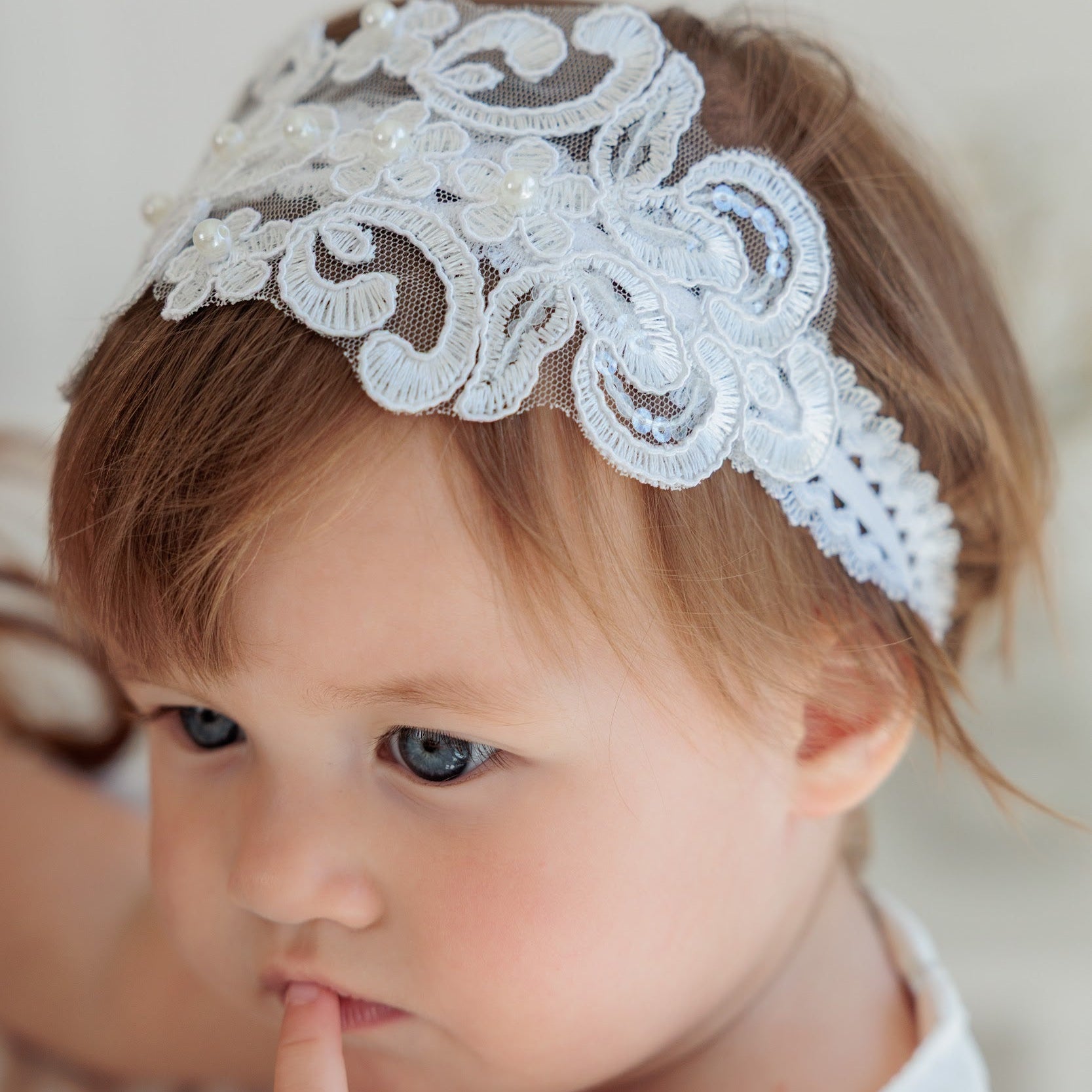 A close-up of a toddler with brown hair wearing a white lace headband with floral embroidery and small pearl-like accents, against a soft light background.