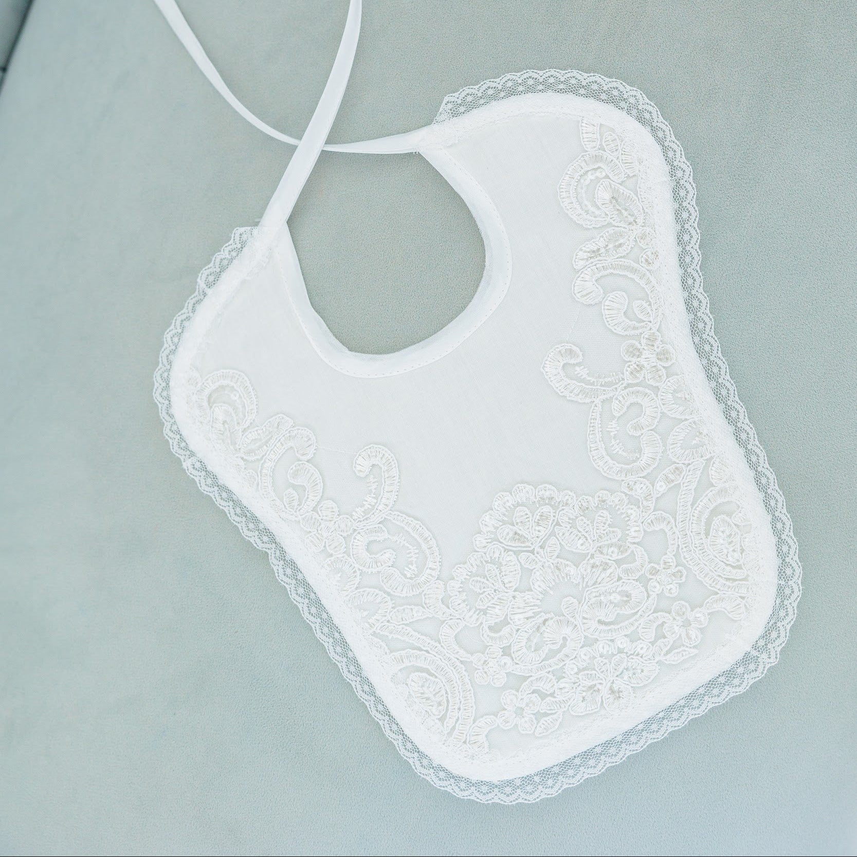 A white baby bib with delicate lace trim and embroidered floral patterns, featuring two thin ties, laid flat on a light sage green fabric surface.