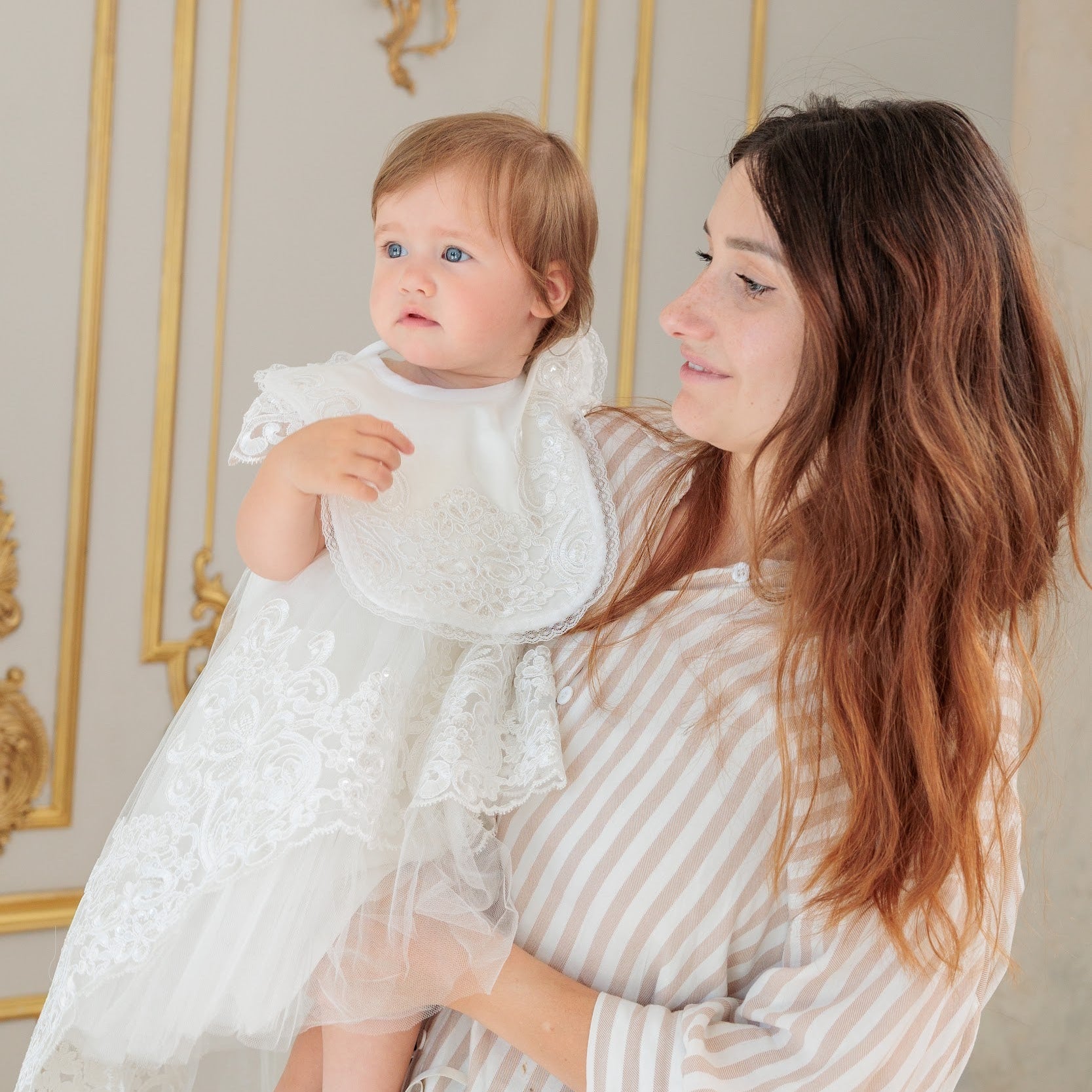 A woman holding a baby girl in a white lace dress, set against an elegant interior with gold-trimmed cream walls.