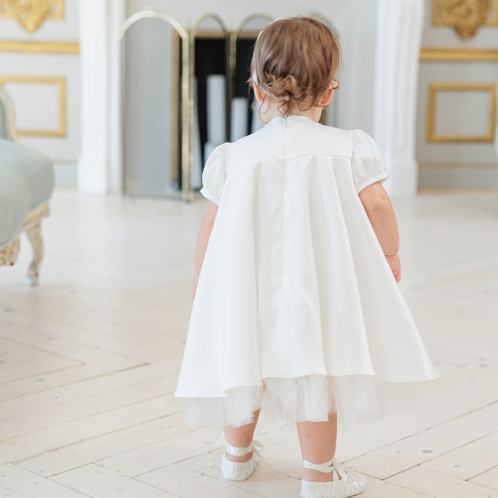 A toddler girl viewed from behind wearing a white short-sleeve dress with a flared cape-like back and a sheer tulle hem, standing in a bright, elegant room with a white fireplace and gold-trimmed walls.