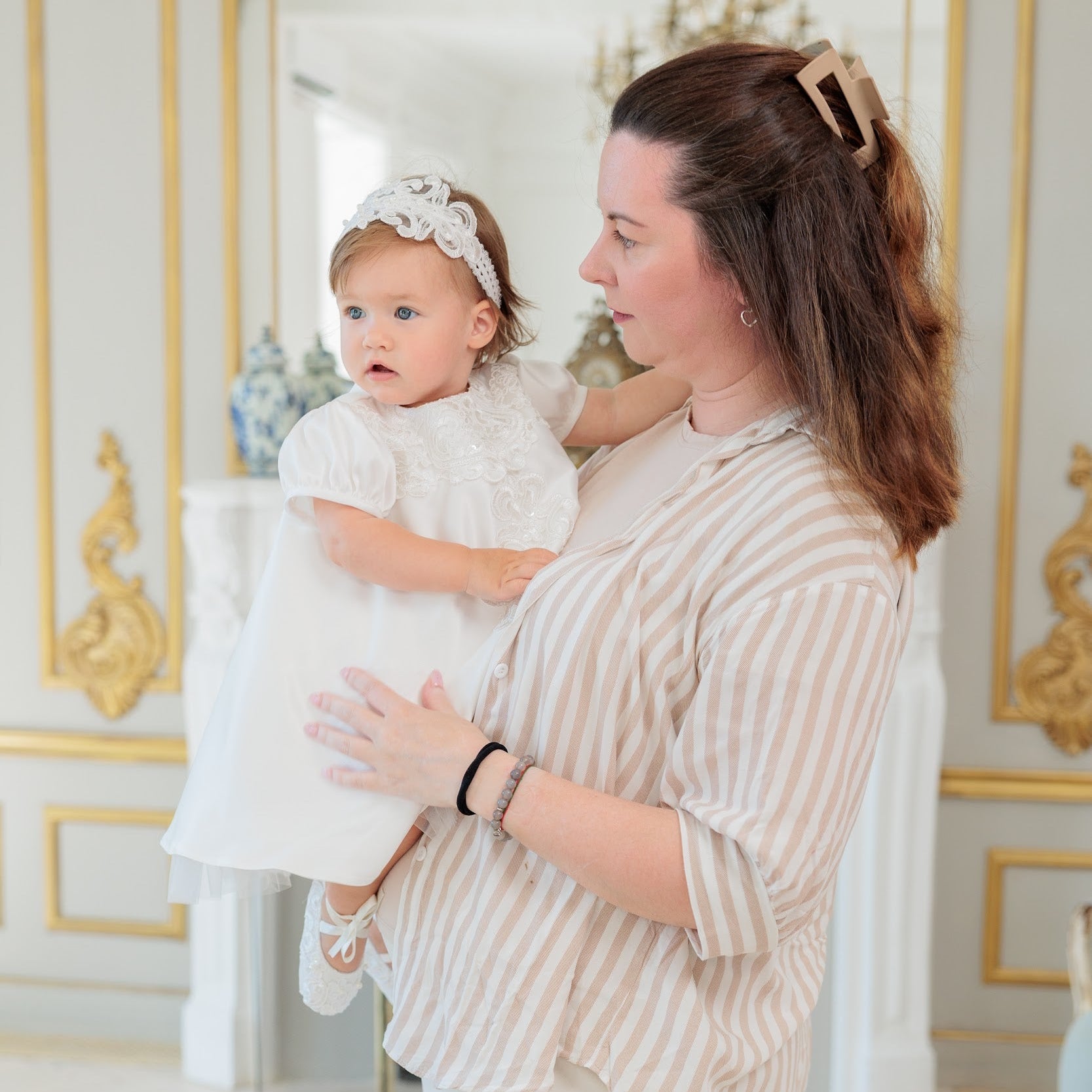 A woman in a beige and white striped shirt holds a baby girl wearing a white baptism dress and matching headband in an elegant room with gold-trimmed white walls.