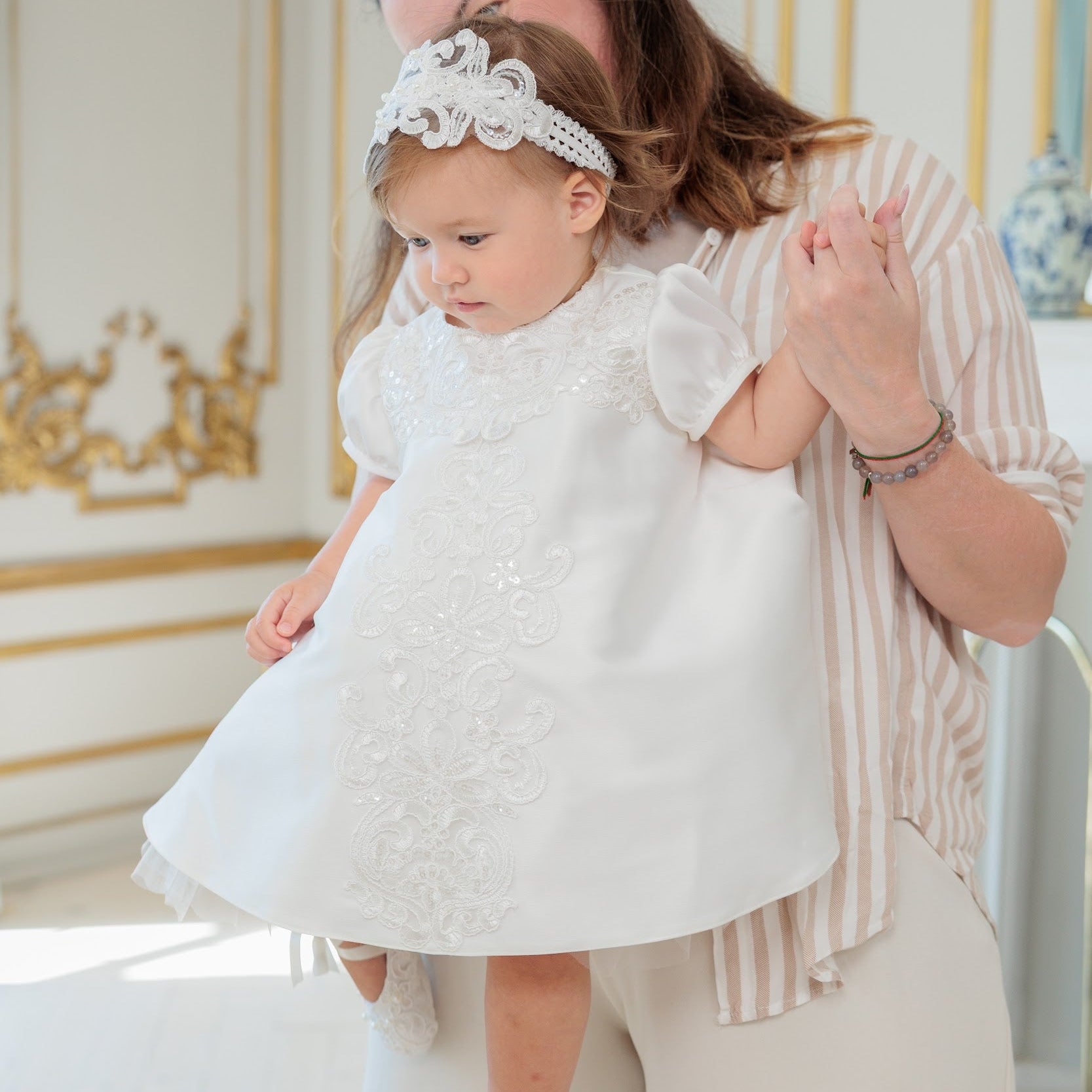 A woman in a cream and pink striped shirt holds a toddler wearing a white baptism dress with lace applique, a white lace headpiece, and white lace shoes in an elegant room with gold-trimmed walls.