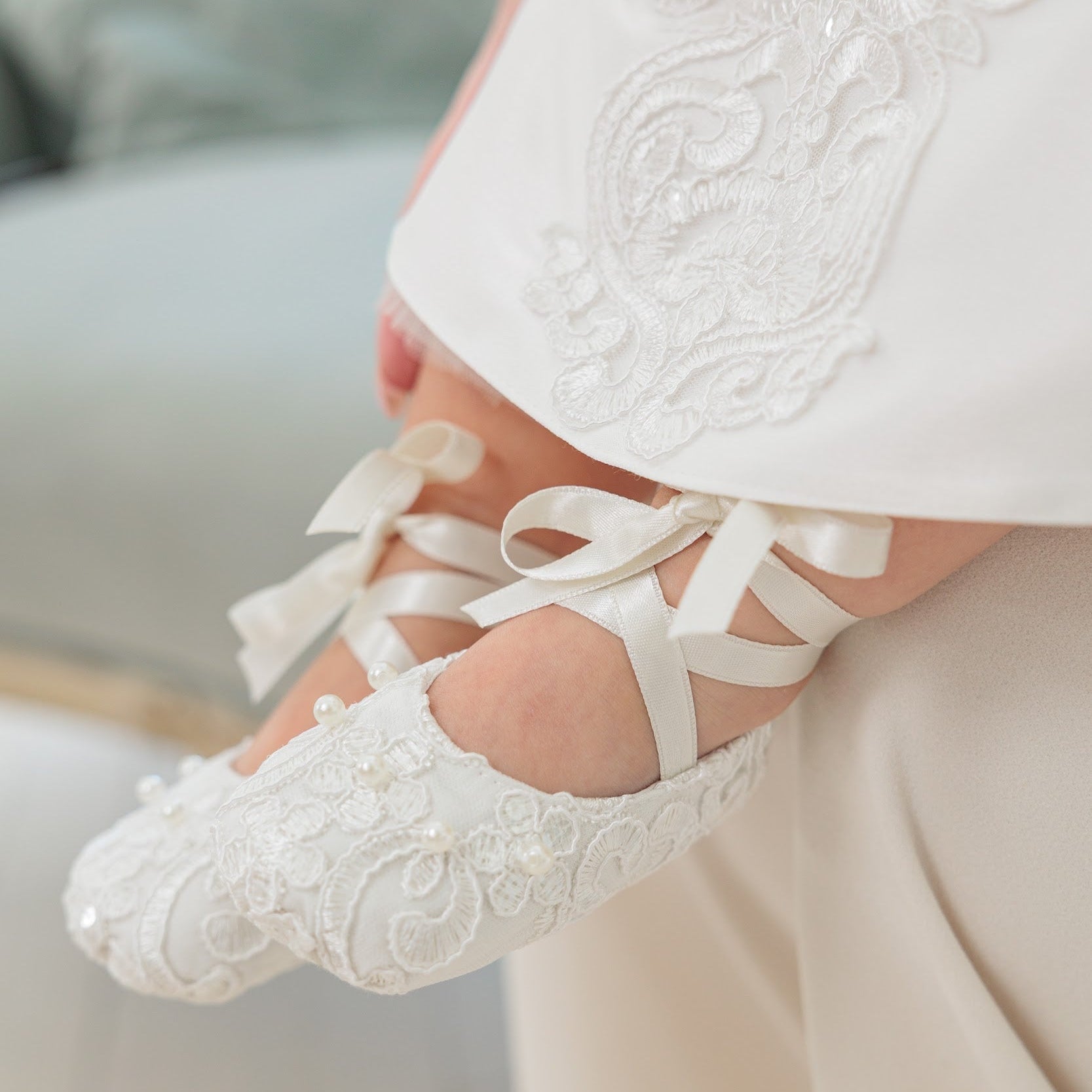 White lace baby shoes with ribbon laces and a matching white embroidered dress, shown on a baby's feet in a soft indoor setting.