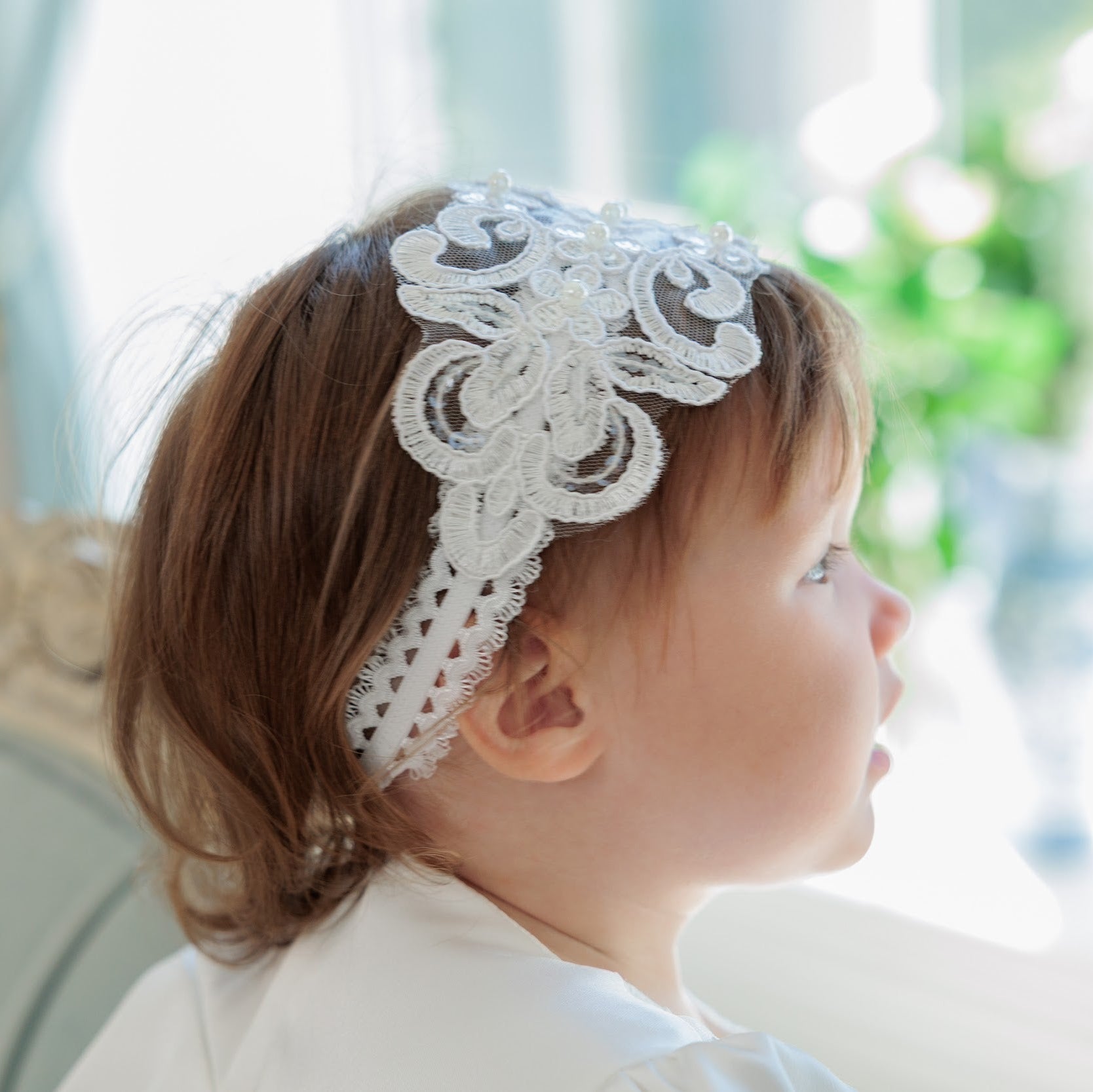 A young child in profile wearing a white lace headband with decorative floral embroidery, set against a bright, softly blurred indoor background.