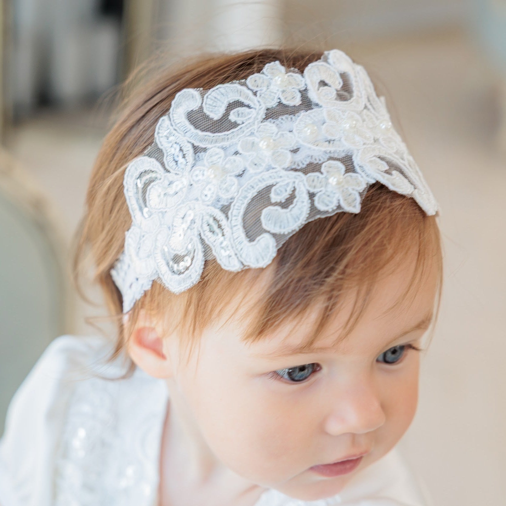 A close-up of a toddler wearing a white lace headband with floral and scroll patterns, set against a softly blurred indoor background.