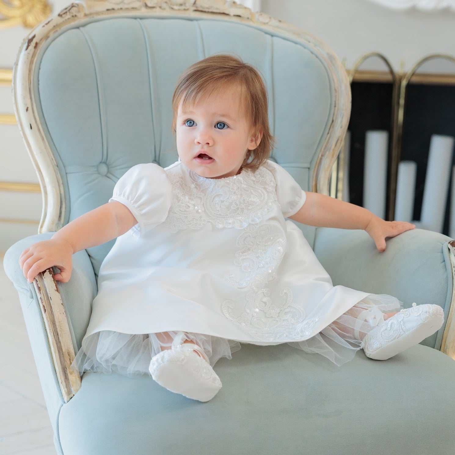 A baby girl in a white embroidered dress and tulle skirt sits on a light blue vintage armchair in front of a white fireplace mantel with gold trim.