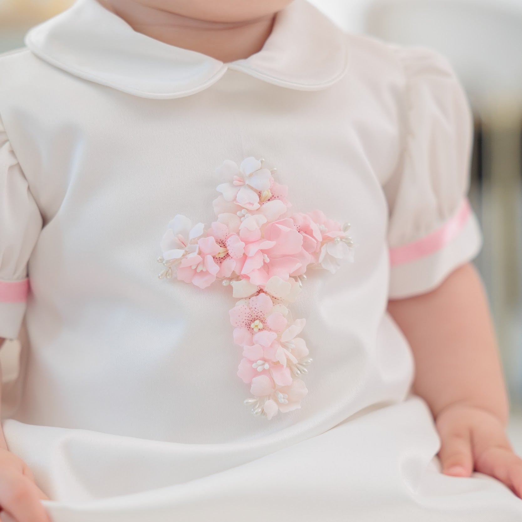 A close-up of a baby wearing a white first communion dress with a pink floral cross applique on the chest, pink trim on the sleeves and hem, and a Peter Pan collar.