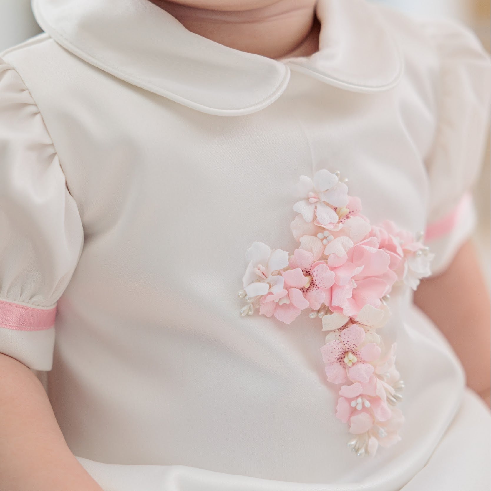 A close-up of a baby wearing a cream-colored short-sleeve dress with a pink band on the sleeve and a pink floral cross applique on the chest.