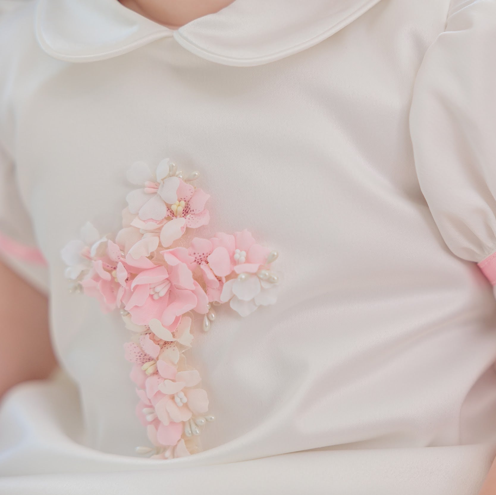 A close-up of a baby wearing a white short-sleeve dress with a pink floral cross applique on the chest and a pink ribbon trim on the sleeve cuff.
