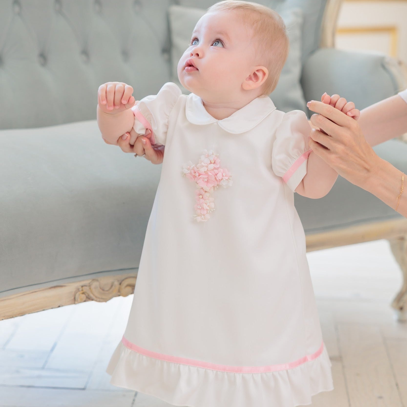 A baby girl wearing a white baptism dress with a pink embroidered cross and pink trim, standing on a light wooden floor and holding an adult's hands for support.