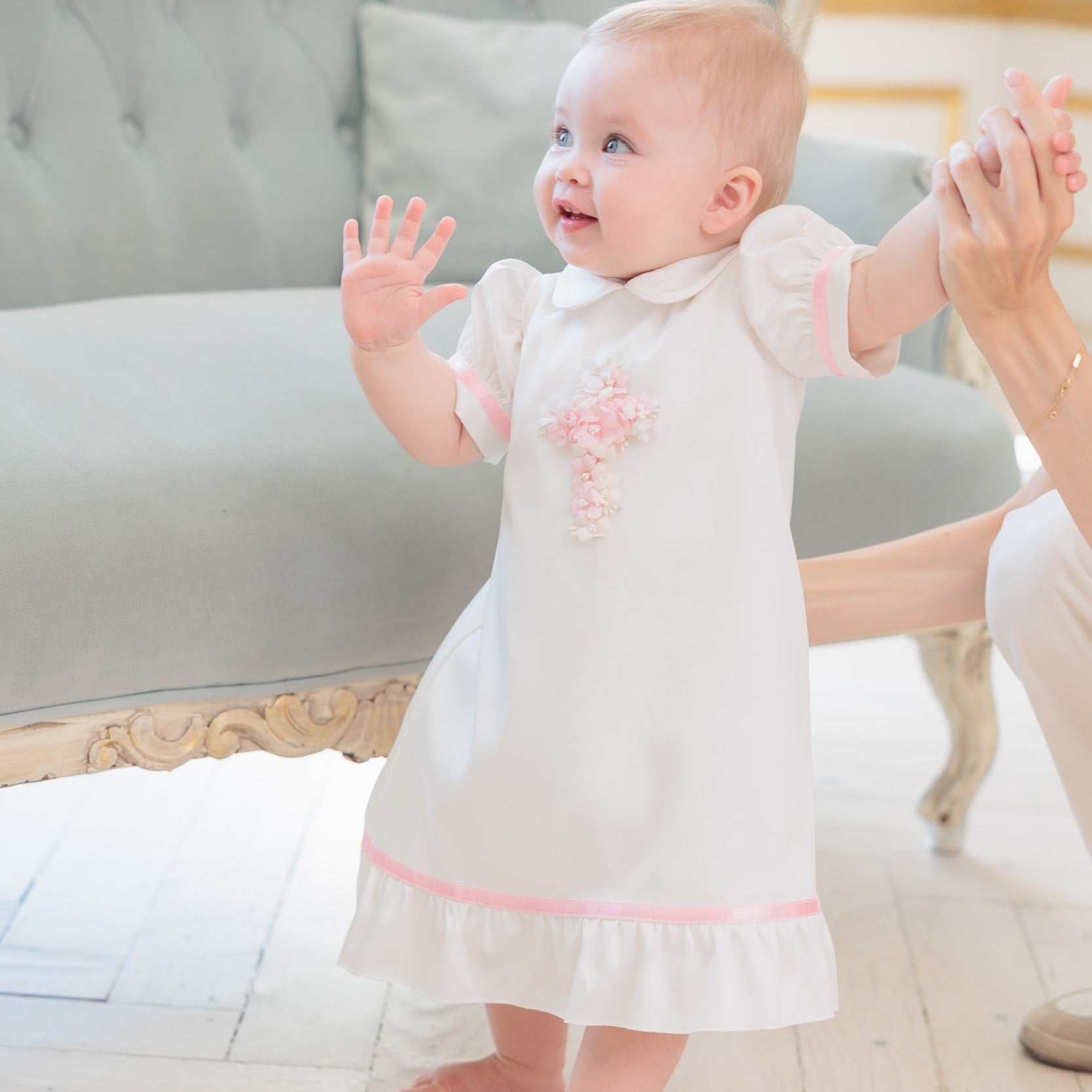 A baby girl wearing a white baptism dress with a pink embroidered cross and pink trim, standing barefoot on a light wooden floor and holding an adult's hand.