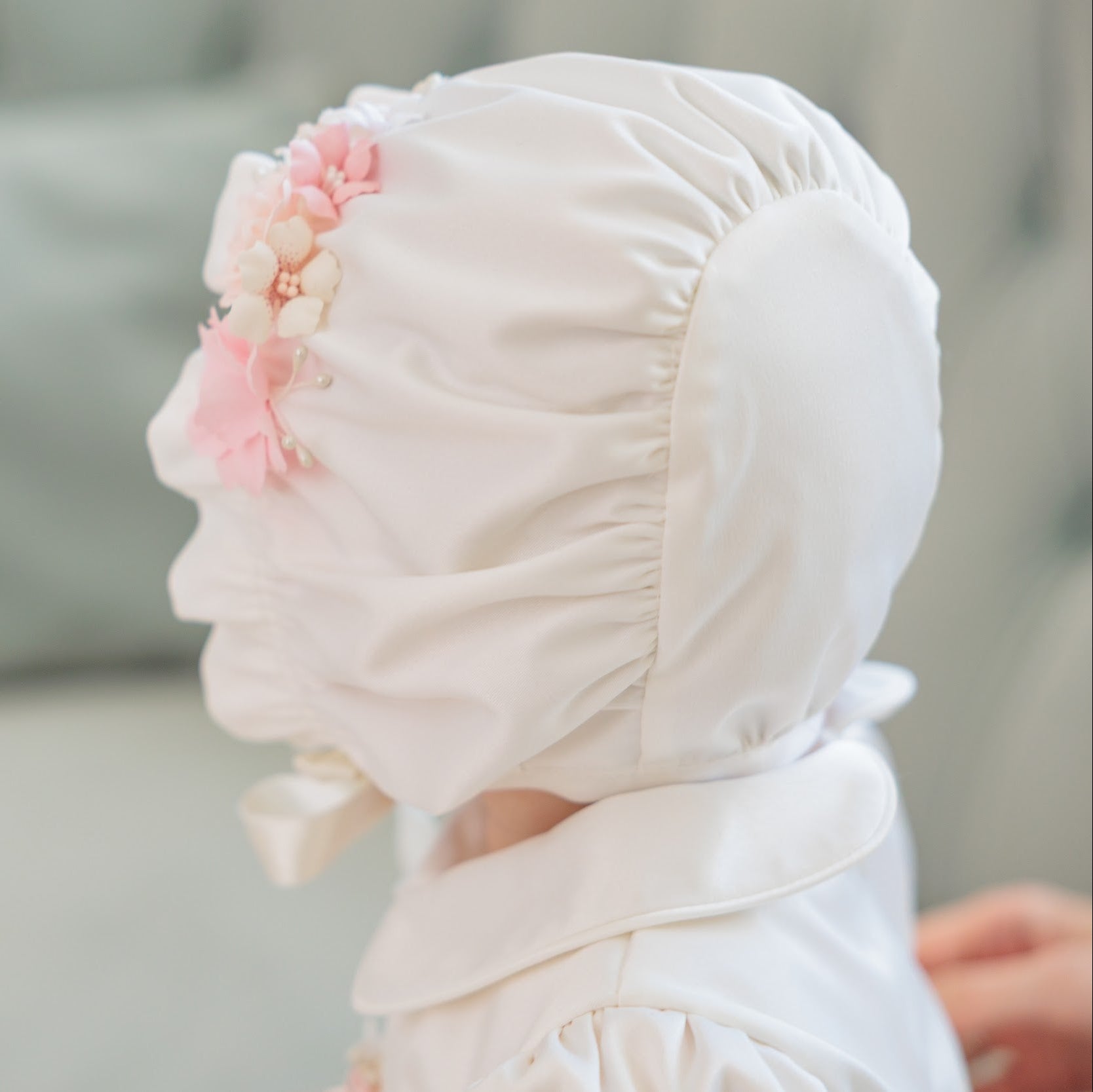 A close-up side profile of a baby wearing a white bonnet with pink and cream fabric flowers, set against a soft, blurred gray background.