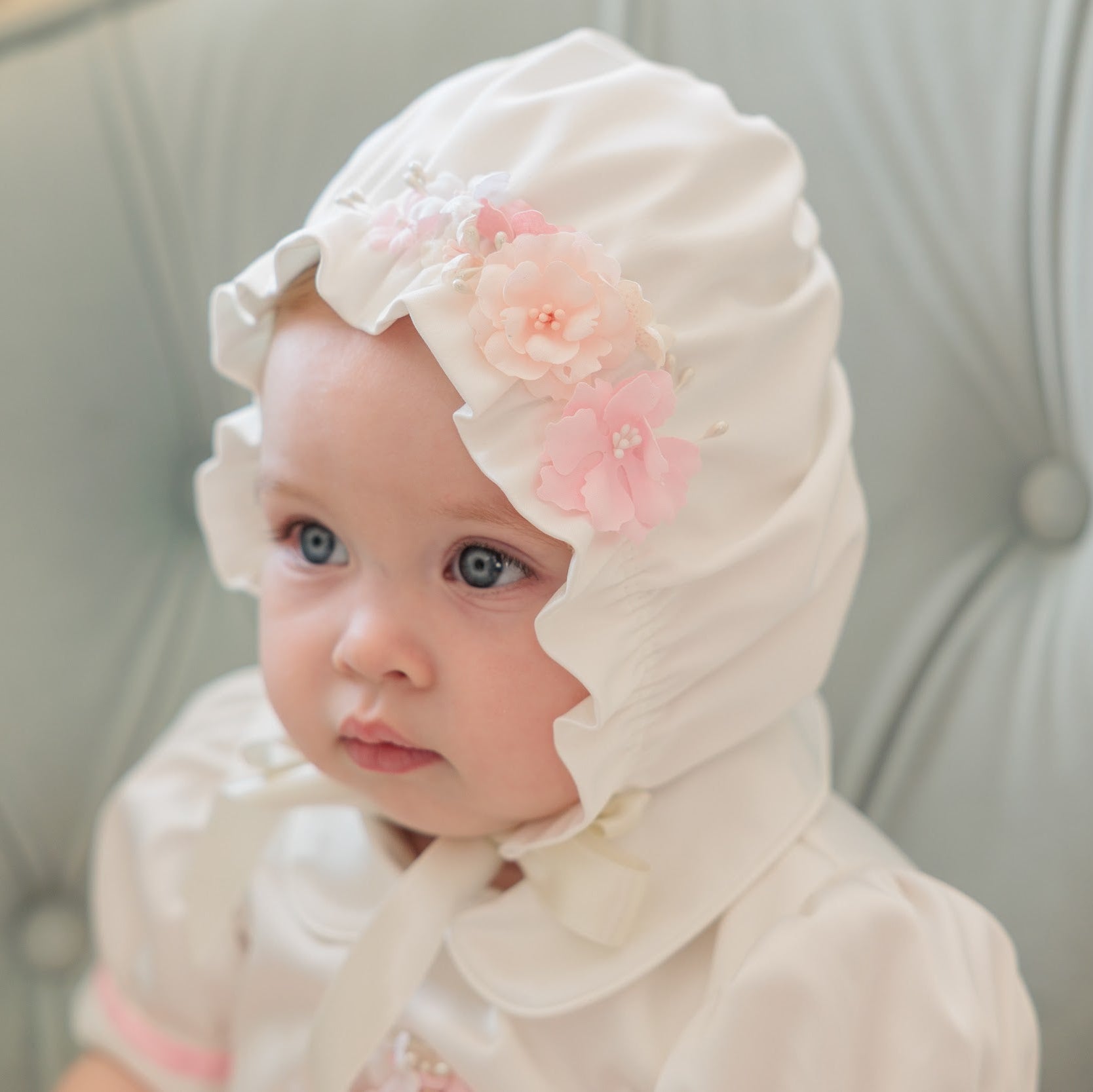 A baby wearing a white bonnet with pink fabric flowers, sitting on a light gray tufted chair.
