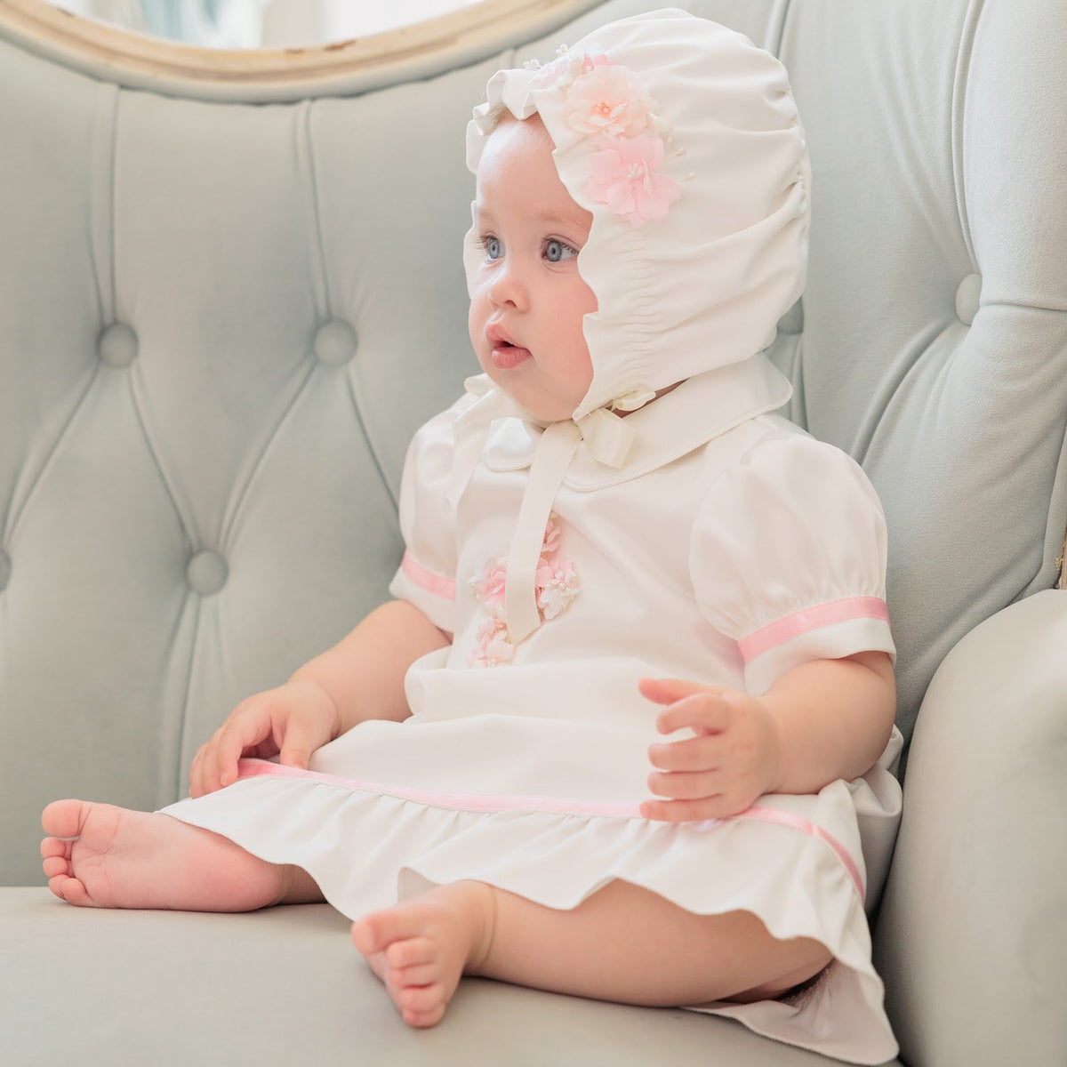 A baby wearing a white bonnet and dress with pink trim sits on a tufted light gray-green armchair.