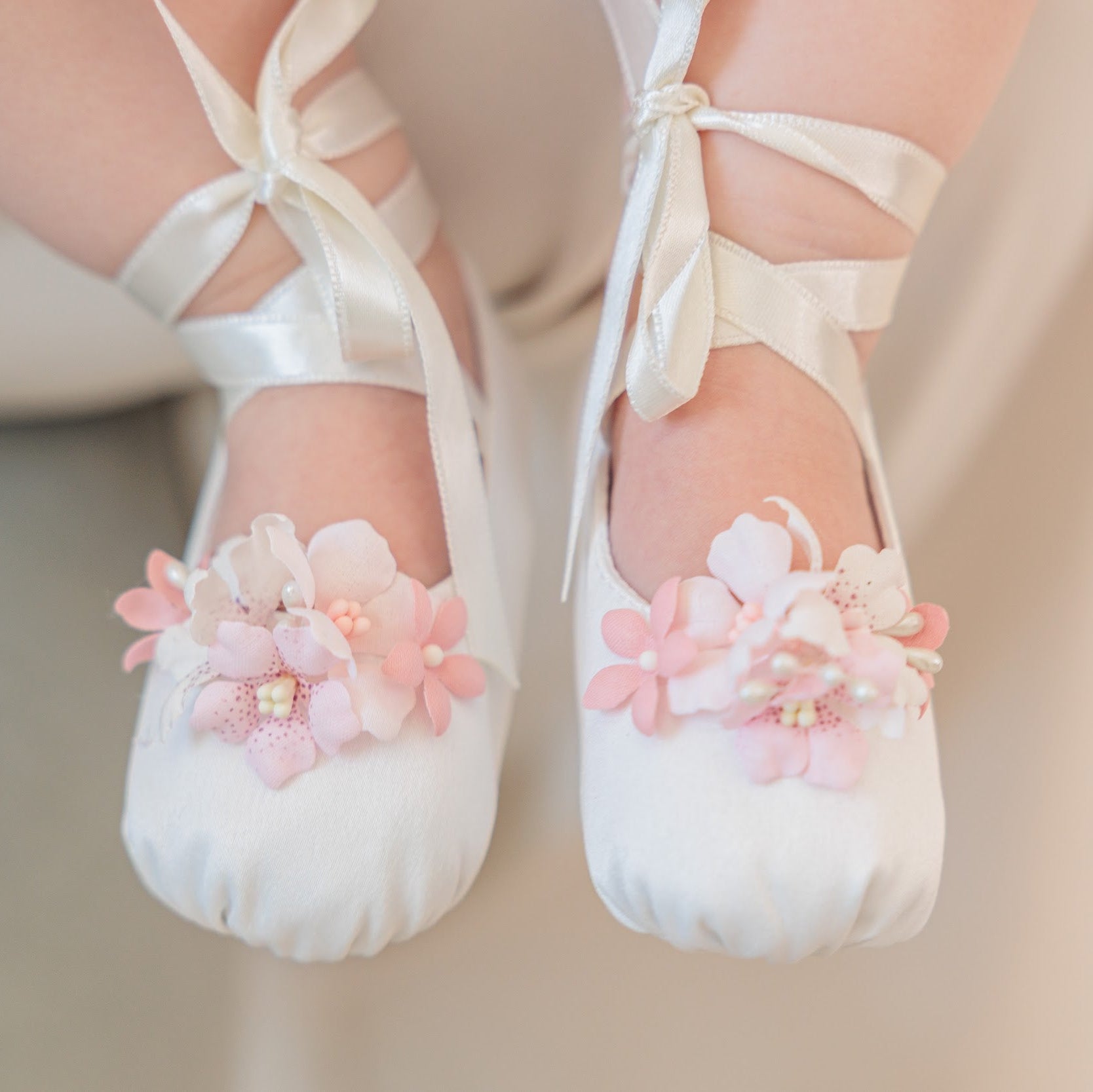 White ballet pointe shoes with pink fabric flowers and pearl accents, worn on a child's feet with white ribbon tights.
