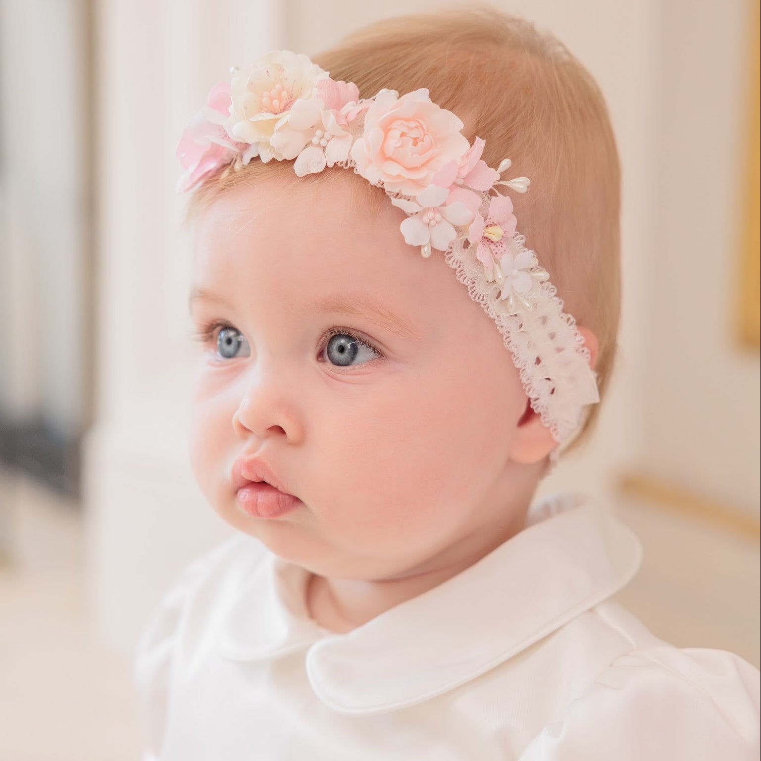 A baby girl with light hair and blue eyes wearing a pink and white floral headband and a white dress with pink accents, looking off-camera in a softly lit indoor setting.