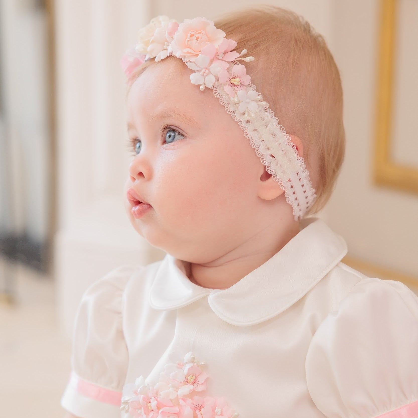 A baby girl wearing a white short-sleeve dress with pink trim and a matching pink and white floral headband, looking off-camera in a softly lit indoor setting.