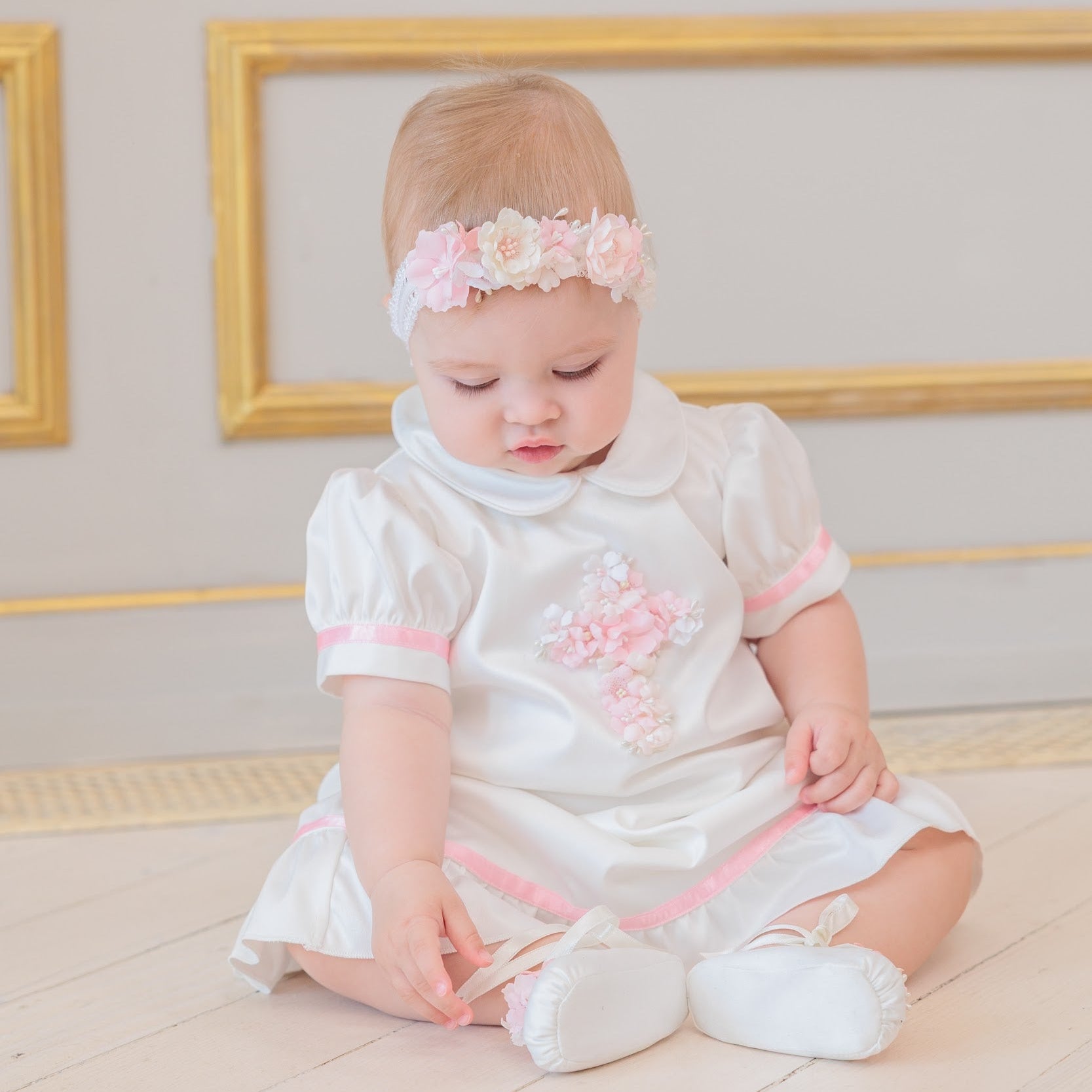 A baby girl sitting on a light wood floor wearing a white satin first communion dress with pink trim, a matching pink and white floral headband, and white shoes.