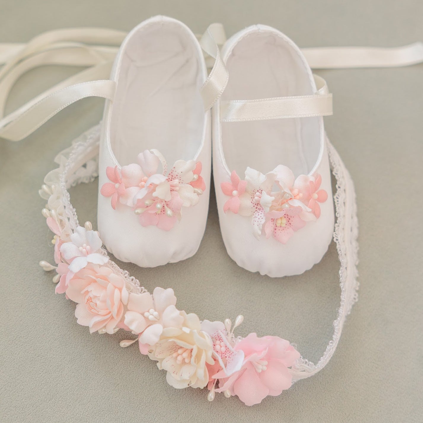 White baby ballet slippers and a matching floral headband with pink and cream fabric flowers, arranged on a soft beige background.
